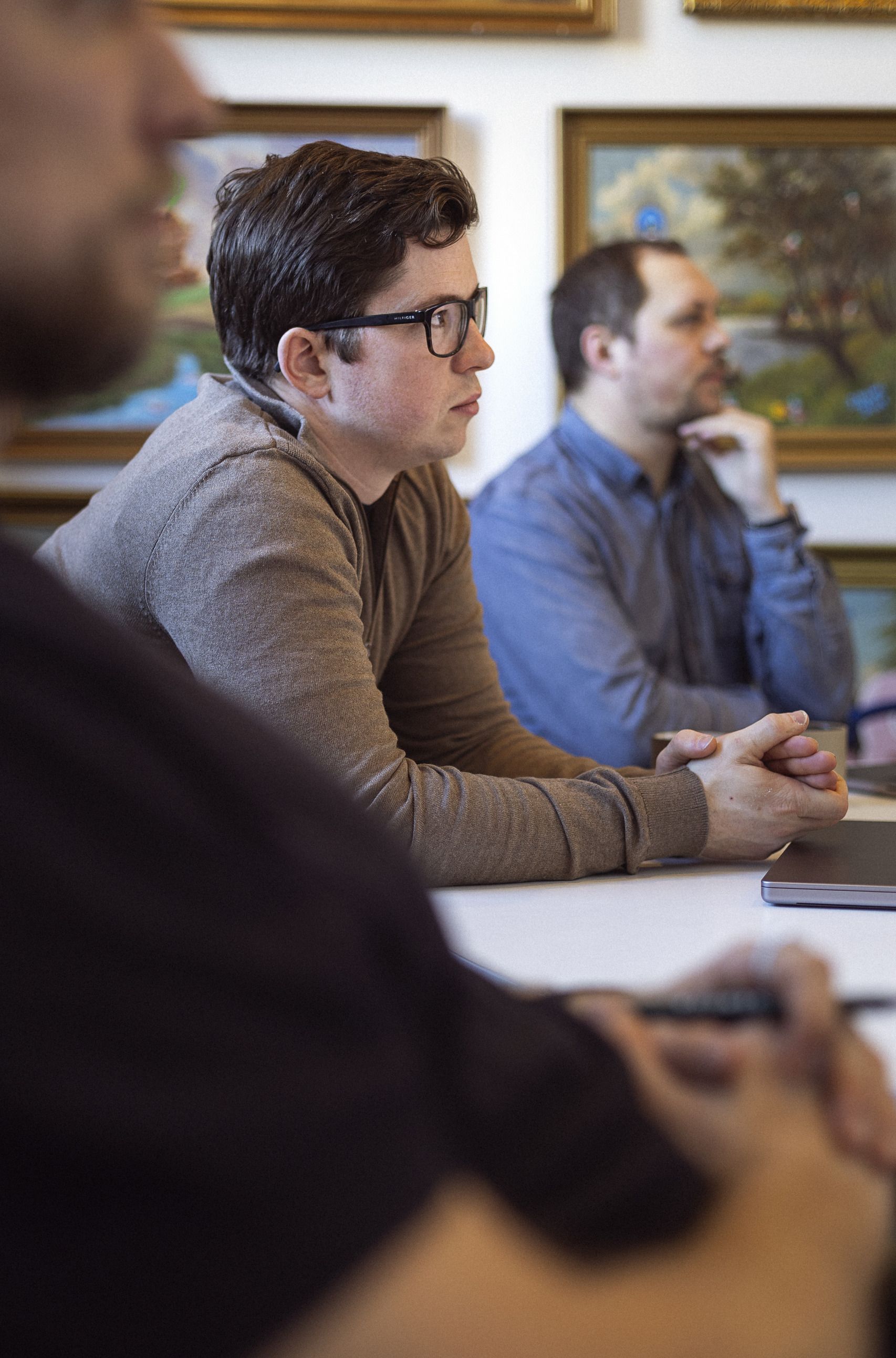 Tre mænd sidder ved et bord og lytter opmærksomt, mens de diskuterer. I baggrunden ses malerier på væggen, og der er fokus på mændenes ansigtsudtryk og hænder, der viser koncentration.