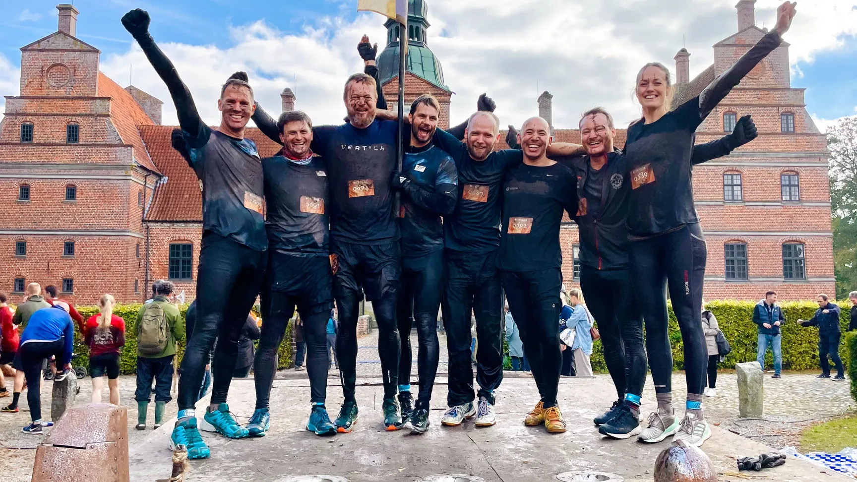 Gruppe af glade deltagere iført sorte trøjer, der står på en platform efter en mudderløb, mens de jubler og strækker armene op i vejret, med en historisk bygning i baggrunden og tilskuere i fokus.