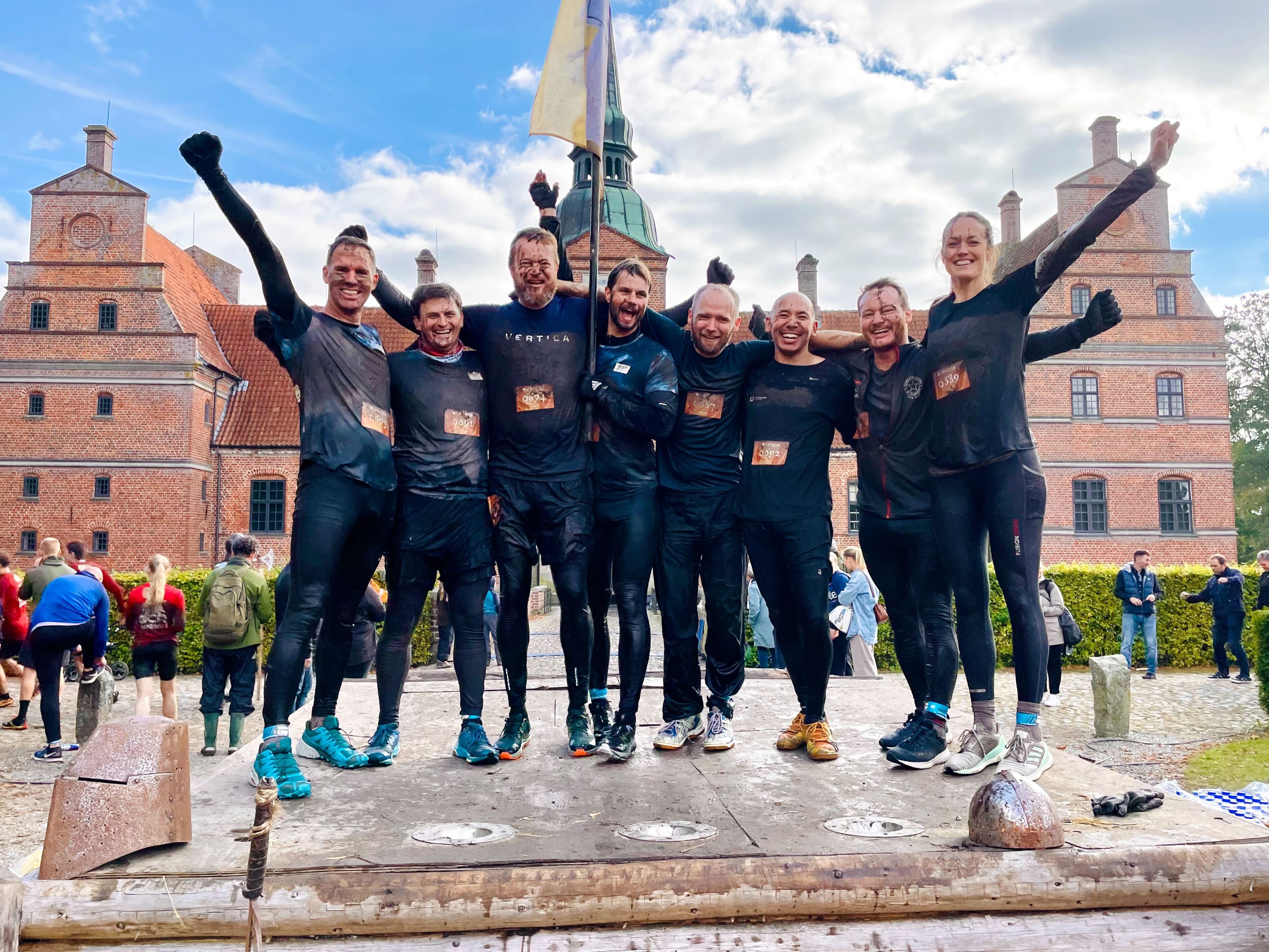 Gruppe af glade deltagere iført sorte trøjer, der står på en platform efter en mudderløb, mens de jubler og strækker armene op i vejret, med en historisk bygning i baggrunden og tilskuere i fokus.