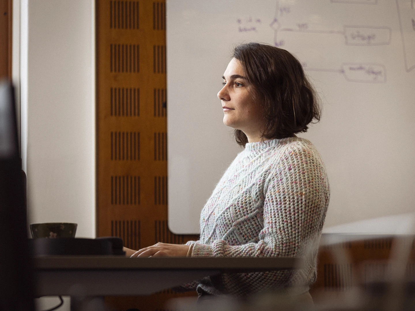 Kvinde med kort hår i en farverig sweater, der sidder foran en computer og ser koncentreret ud, i et lyst rum med en tavle i baggrunden.