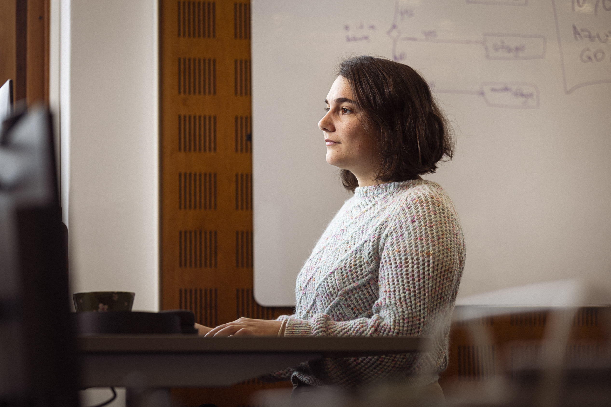Kvinde med kort hår i en farverig sweater, der sidder foran en computer og ser koncentreret ud, i et lyst rum med en tavle i baggrunden.