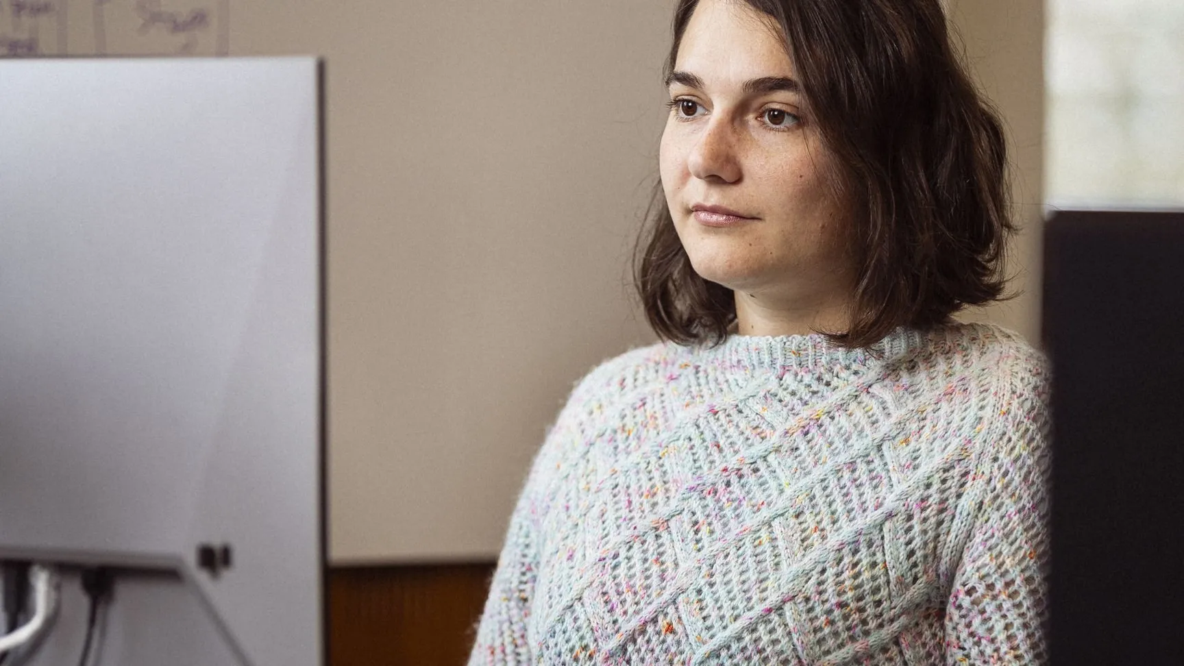 Ung kvinde i farverig striktrøje arbejder koncentreret ved computerbord med en grøn kop og kontorartikler i baggrunden.