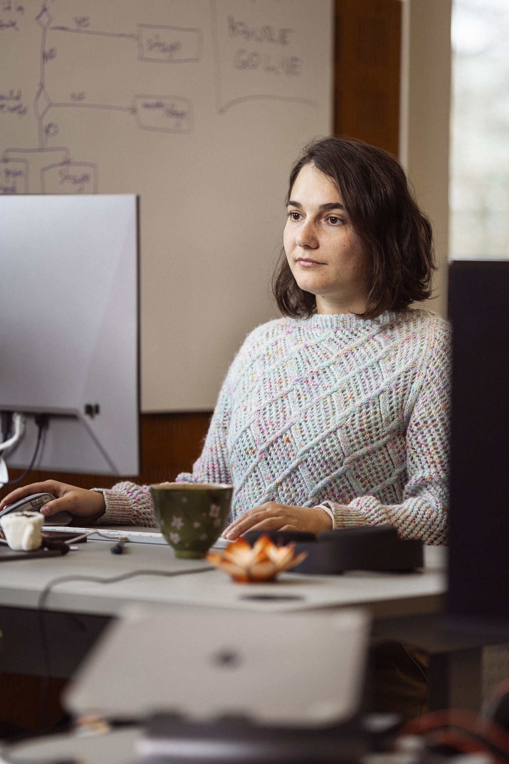 Ung kvinde i farverig striktrøje arbejder koncentreret ved computerbord med en grøn kop og kontorartikler i baggrunden.