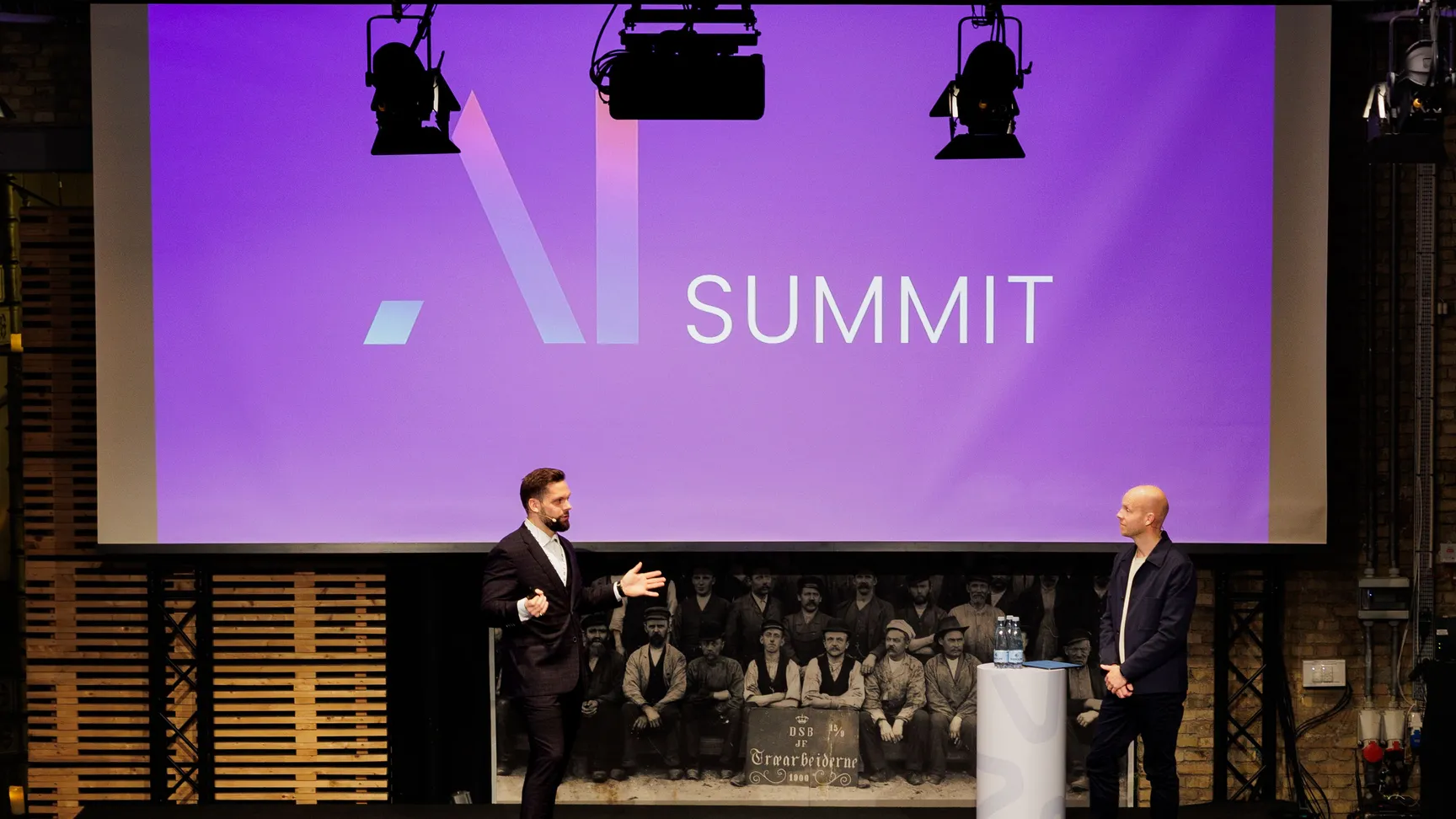 To personer præsenterer ved AI Summit på en scenen foran et publikum, der sidder ved runde borde. Baggrunden viser en stor skærm med teksten "AI SUMMIT" i moderne design.