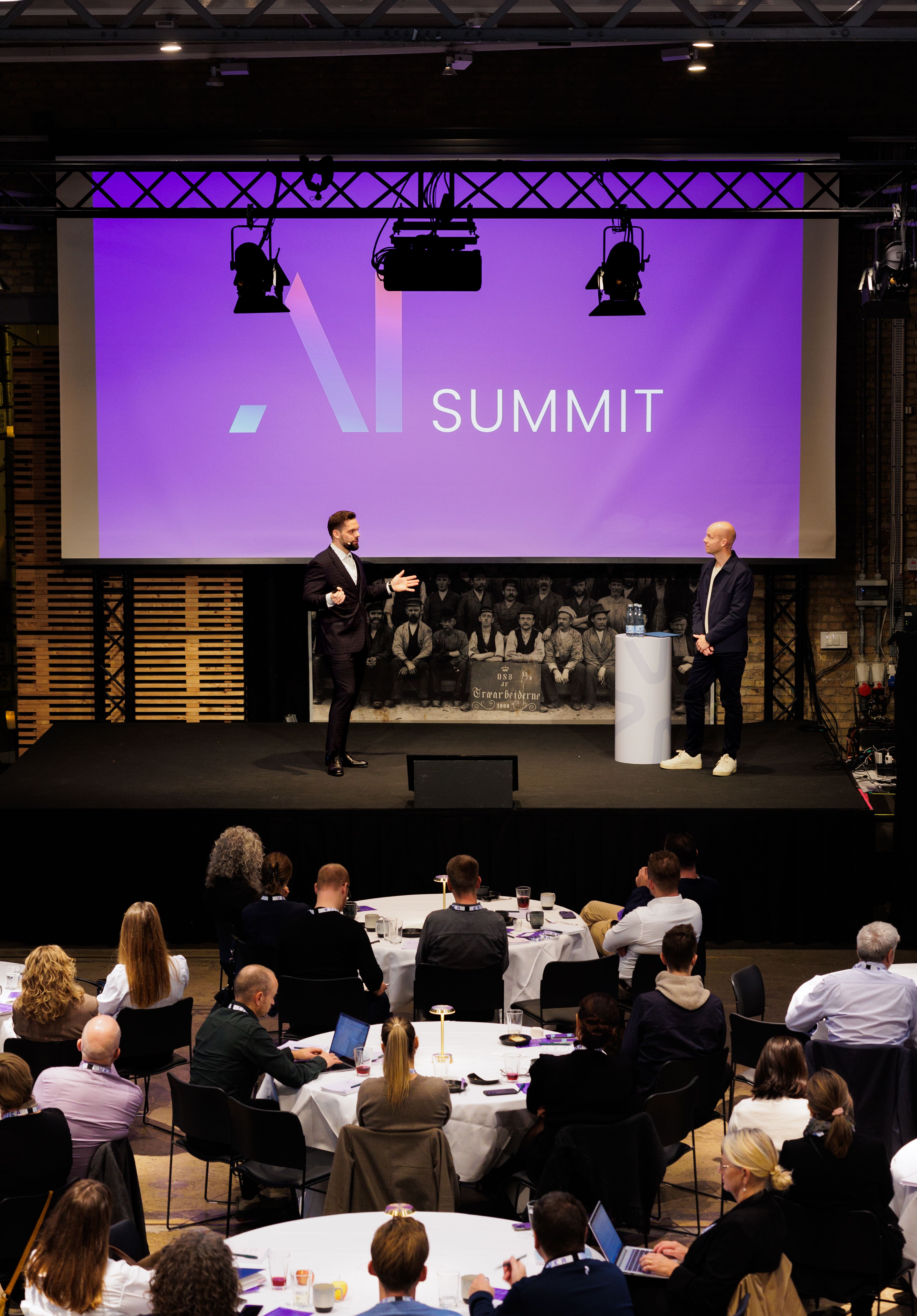 To personer præsenterer ved AI Summit på en scenen foran et publikum, der sidder ved runde borde. Baggrunden viser en stor skærm med teksten "AI SUMMIT" i moderne design.