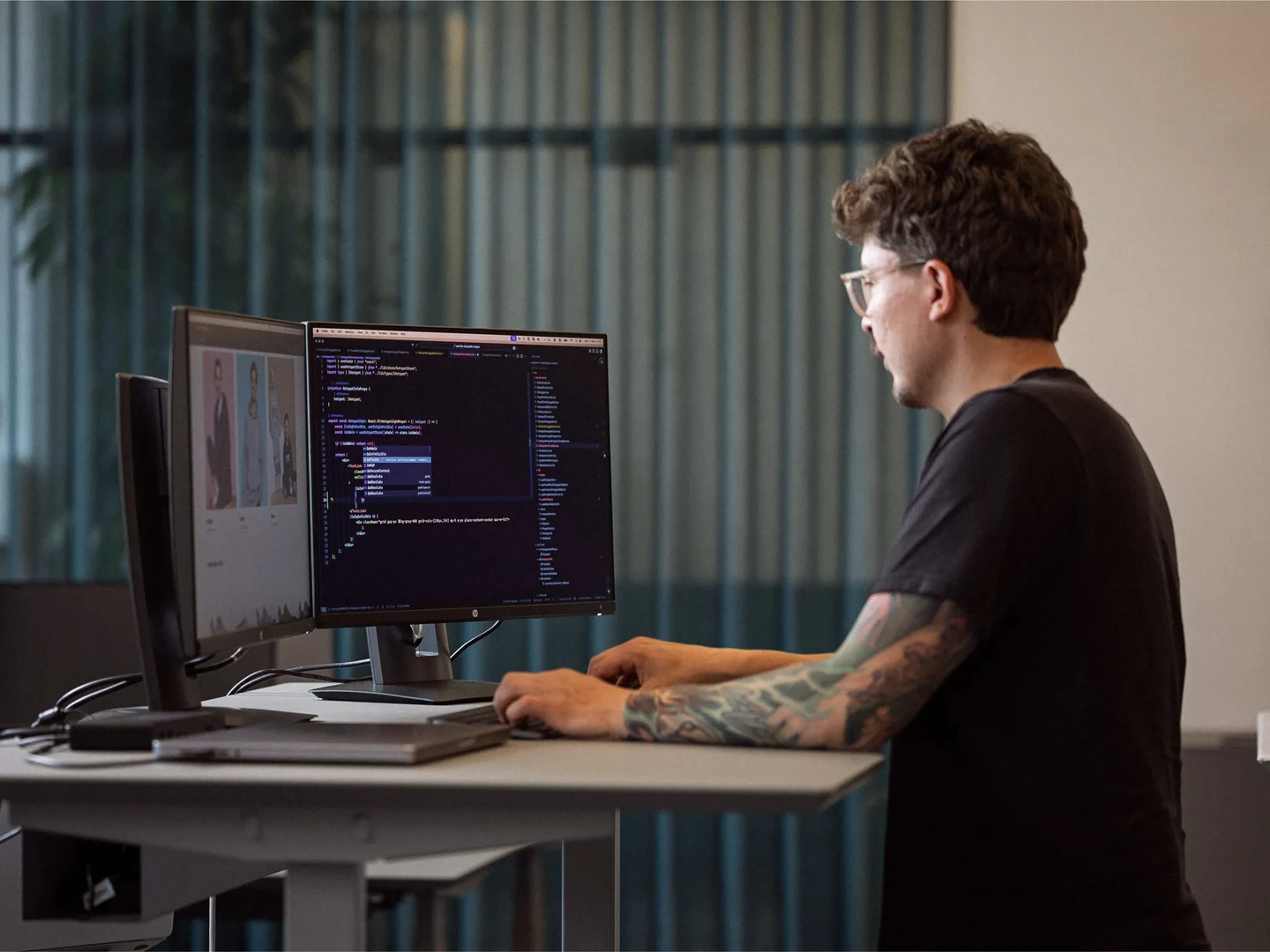 En person med tatoveringer sidder og arbejder ved et skrivebord foran to computermonitorer, hvor den ene viser kode, mens den anden skærm viser billeder. Der er grønne planter og et vindue med gennemsigtige gardiner i baggrunden.