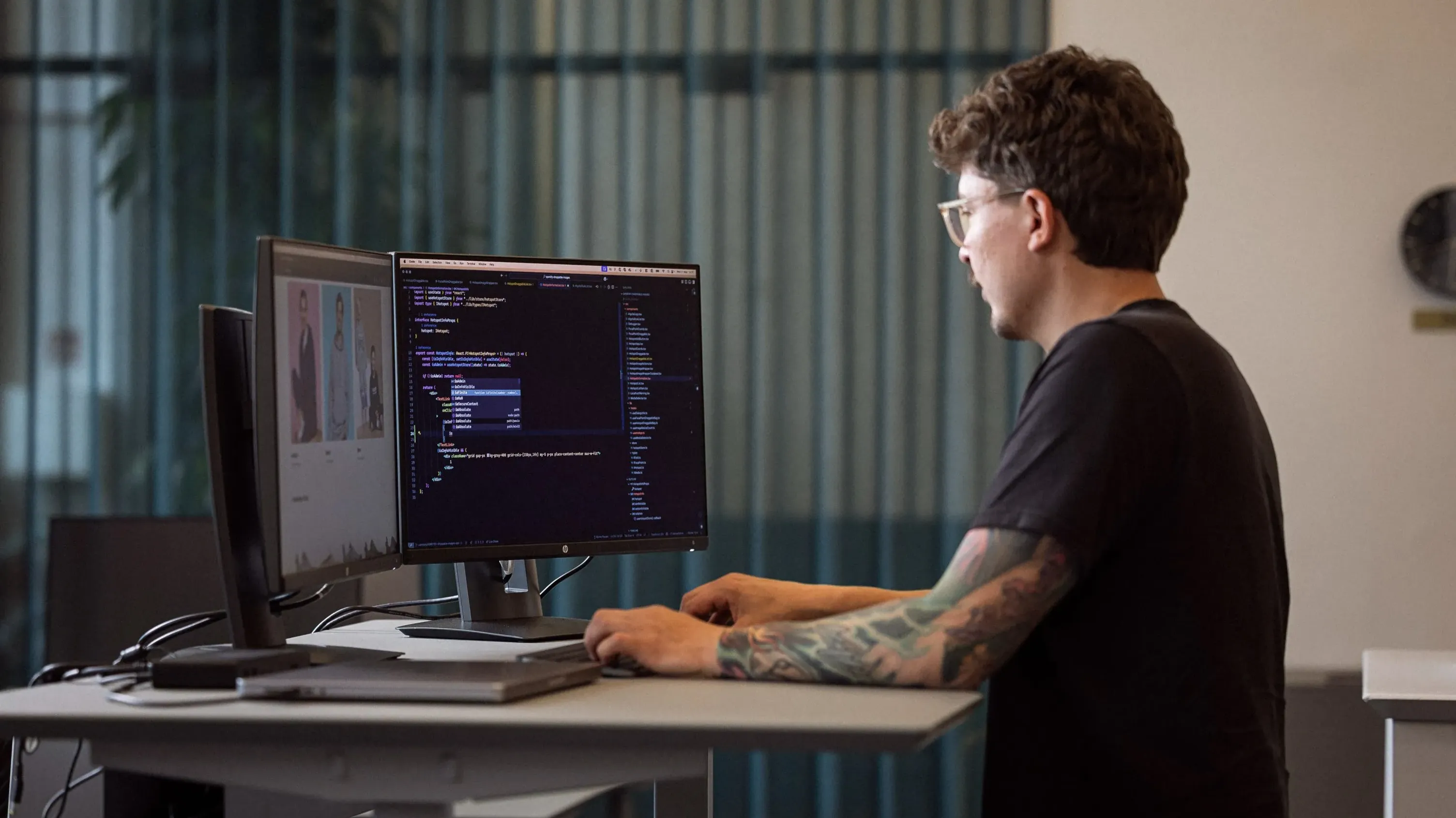 En person med tatoveringer sidder og arbejder ved et skrivebord foran to computermonitorer, hvor den ene viser kode, mens den anden skærm viser billeder. Der er grønne planter og et vindue med gennemsigtige gardiner i baggrunden.