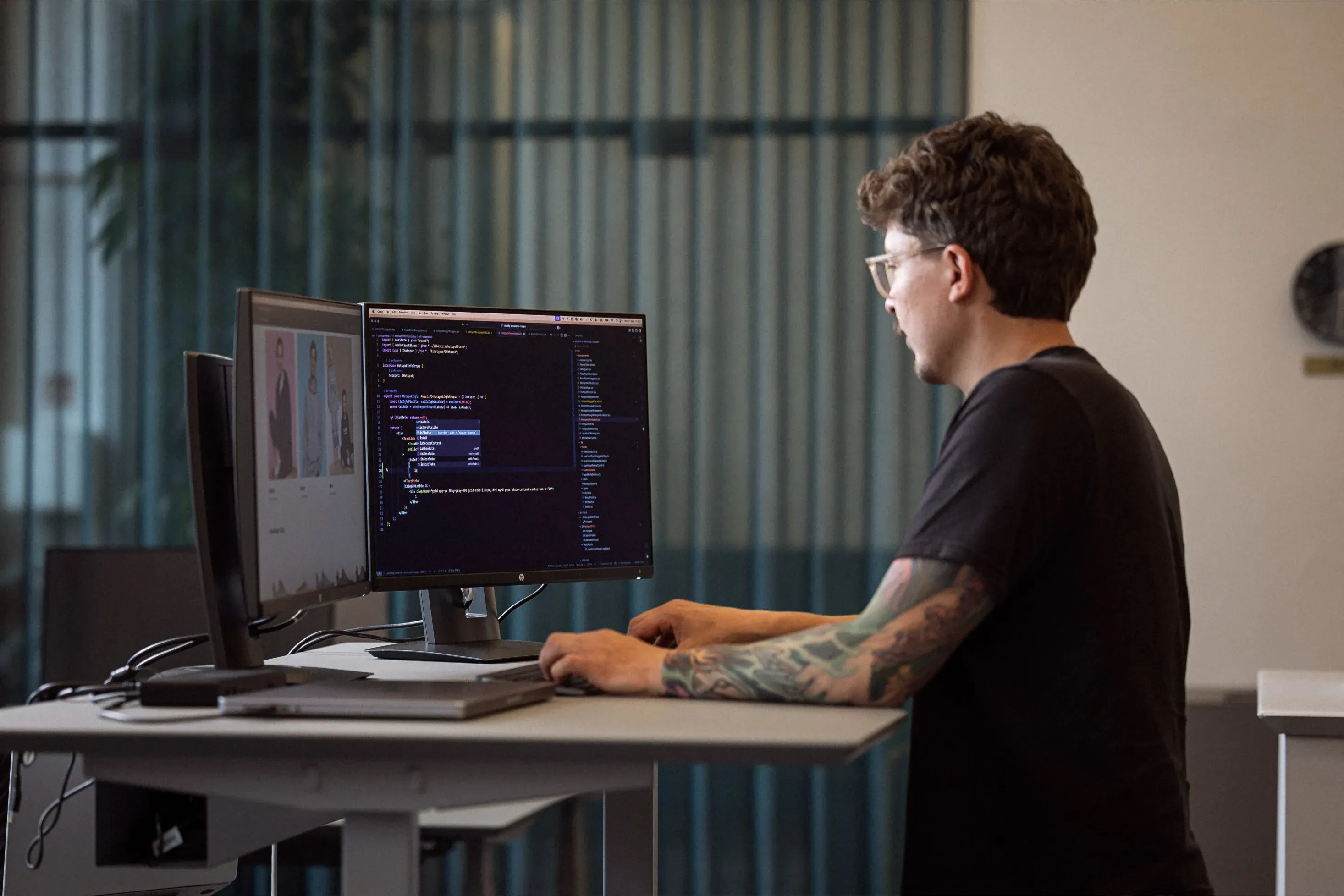 En person med tatoveringer sidder og arbejder ved et skrivebord foran to computermonitorer, hvor den ene viser kode, mens den anden skærm viser billeder. Der er grønne planter og et vindue med gennemsigtige gardiner i baggrunden.