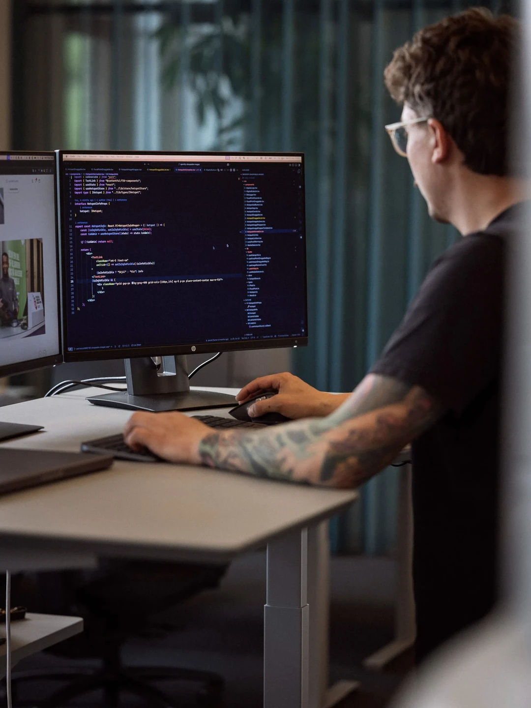 En programmerer arbejder ved en computer med to skærme, hvor den ene viser kode. Personen har tatoveringer på armen og bruger et tastatur. Baggrunden har grønne gardiner.