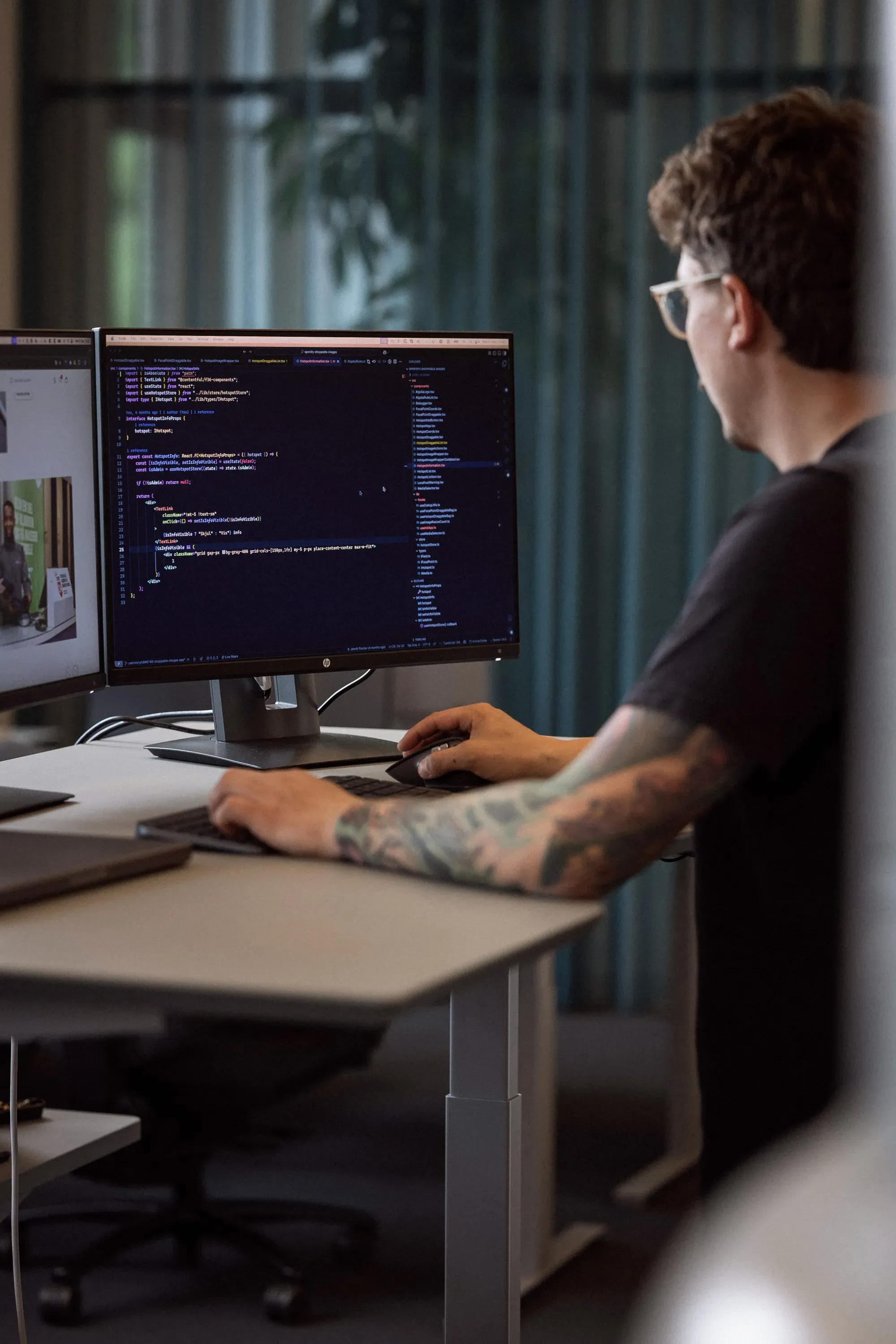 En programmerer arbejder ved en computer med to skærme, hvor den ene viser kode. Personen har tatoveringer på armen og bruger et tastatur. Baggrunden har grønne gardiner.