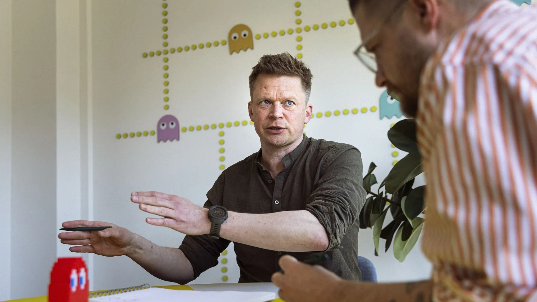 To personer i en samtale ved et bord, med en rød Lego-figur i forsædet og en grøn plante i baggrunden. Den ene person gestikulerer med hånden, mens den anden lytter. Væggen er dekoreret med farverige spøgelsesfigurer og prikker.