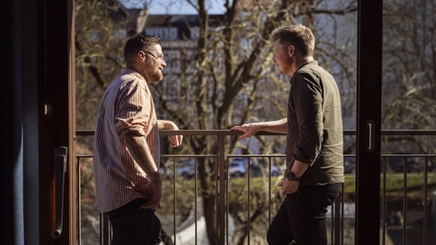 Two men engaged in conversation on a balcony, with a sunny outdoor view featuring trees and buildings in the background.
