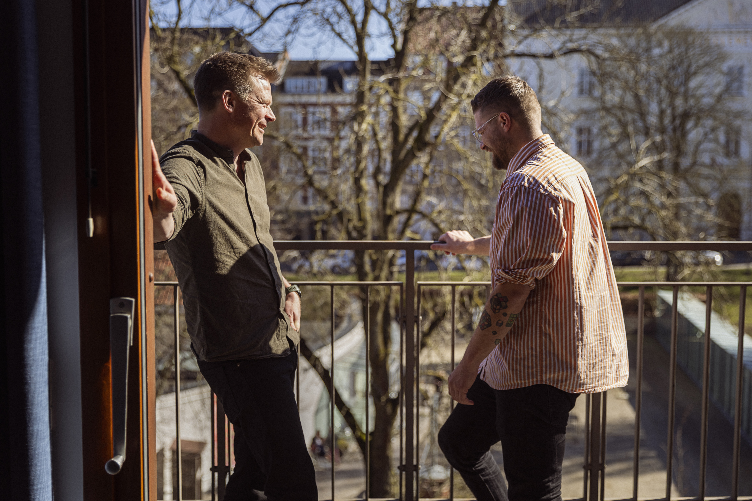 To mænd står på en balkon og griner sammen, omgivet af et grønt træ og bygninger i baggrunden i sollys.