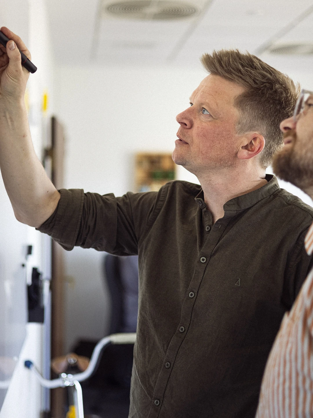 To personer i et kontormiljø står ved et whiteboard, hvor den ene person skriver med en tusch, mens den anden ser opmærksomt på.