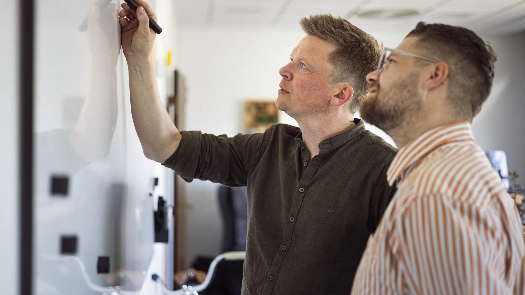 To personer i et kontormiljø står ved et whiteboard, hvor den ene person skriver med en tusch, mens den anden ser opmærksomt på.