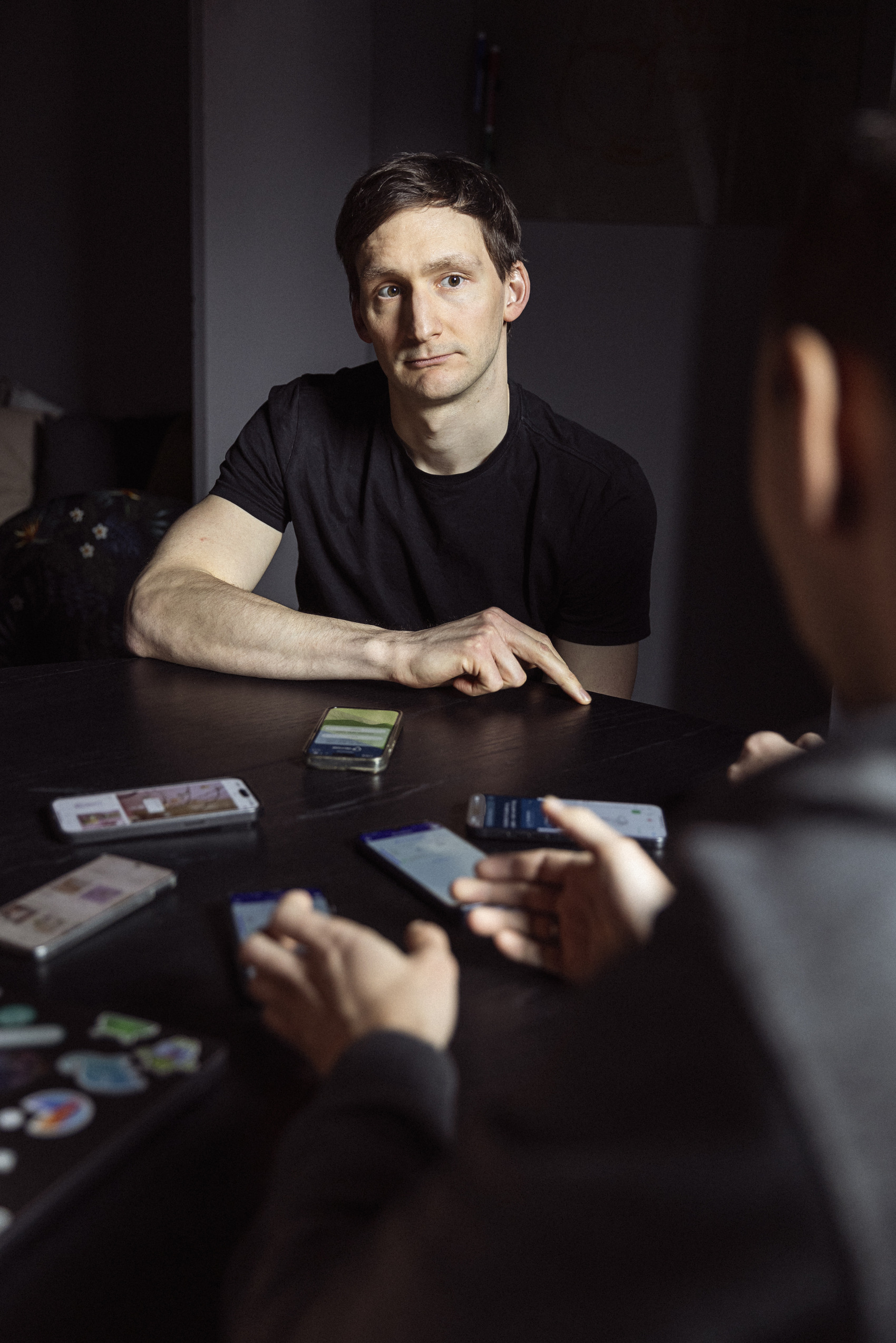 En mand med kort brunt hår sidder ved et bord med flere smartphones, ser eftertænksom ud, mens han kigger mod en person, der ikke er synlig i billedet.