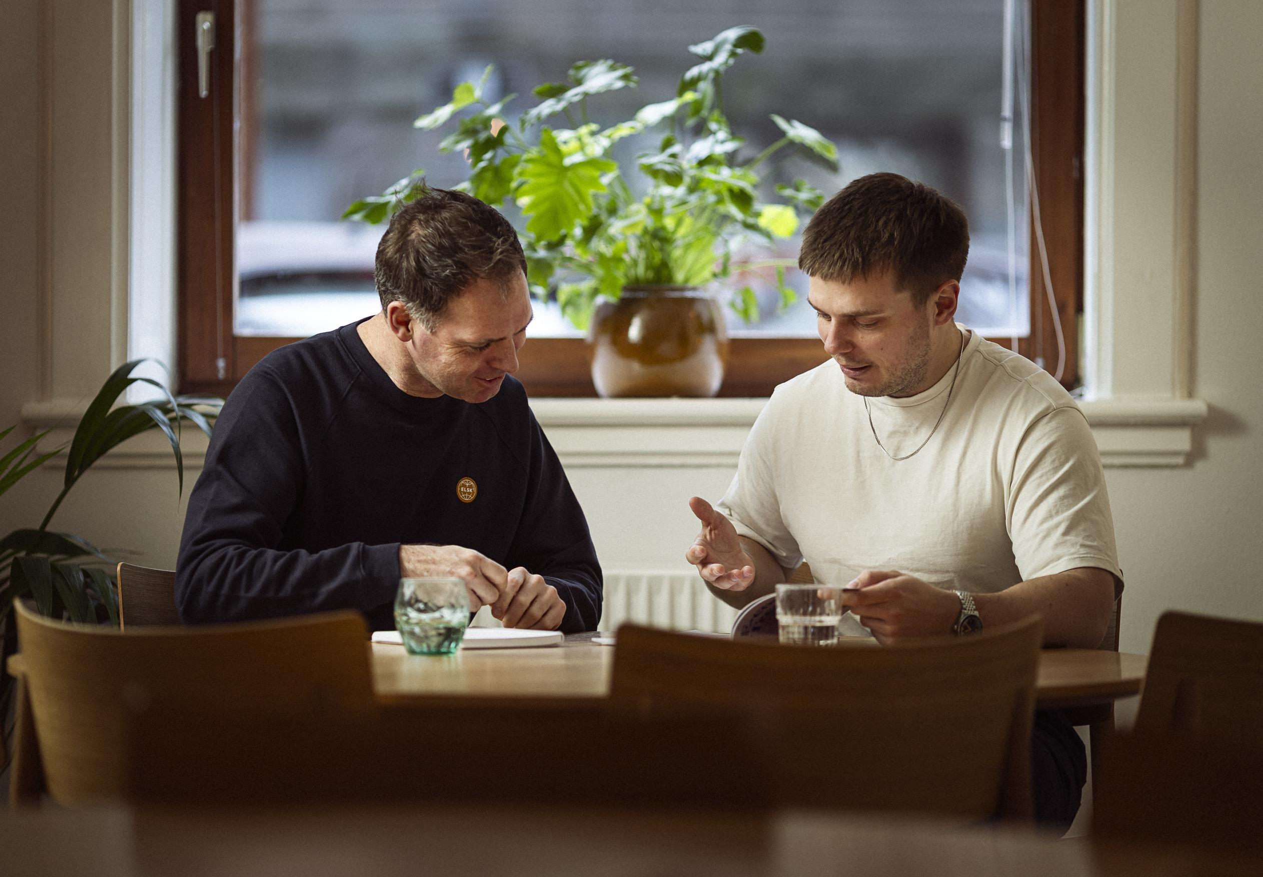 To mænd sidder ved et træbord i en lys café, mens de samtaler og gennemgår noter, omkranset af grønne planter i vindueskarme.