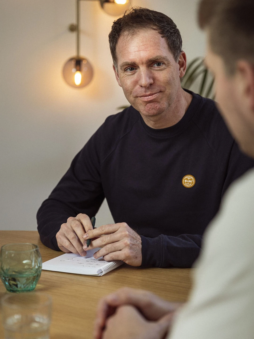 To mænd sidder ved et træbord, hvoraf den ene skriver i en notesblok, mens den anden lytter opmærksomt. På bordet står et glas vand og en grønt krystalglas. Belysningen i rummet er varm og indbydende.