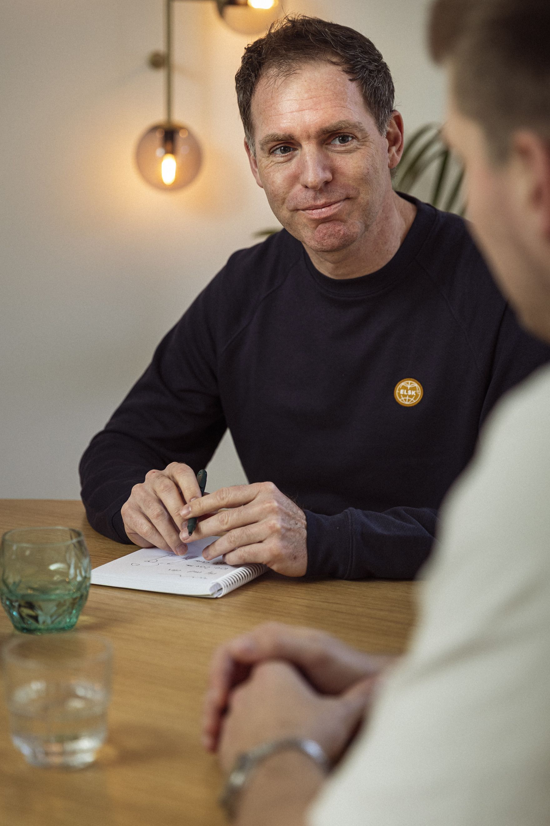 To mænd sidder ved et træbord, hvoraf den ene skriver i en notesblok, mens den anden lytter opmærksomt. På bordet står et glas vand og en grønt krystalglas. Belysningen i rummet er varm og indbydende.