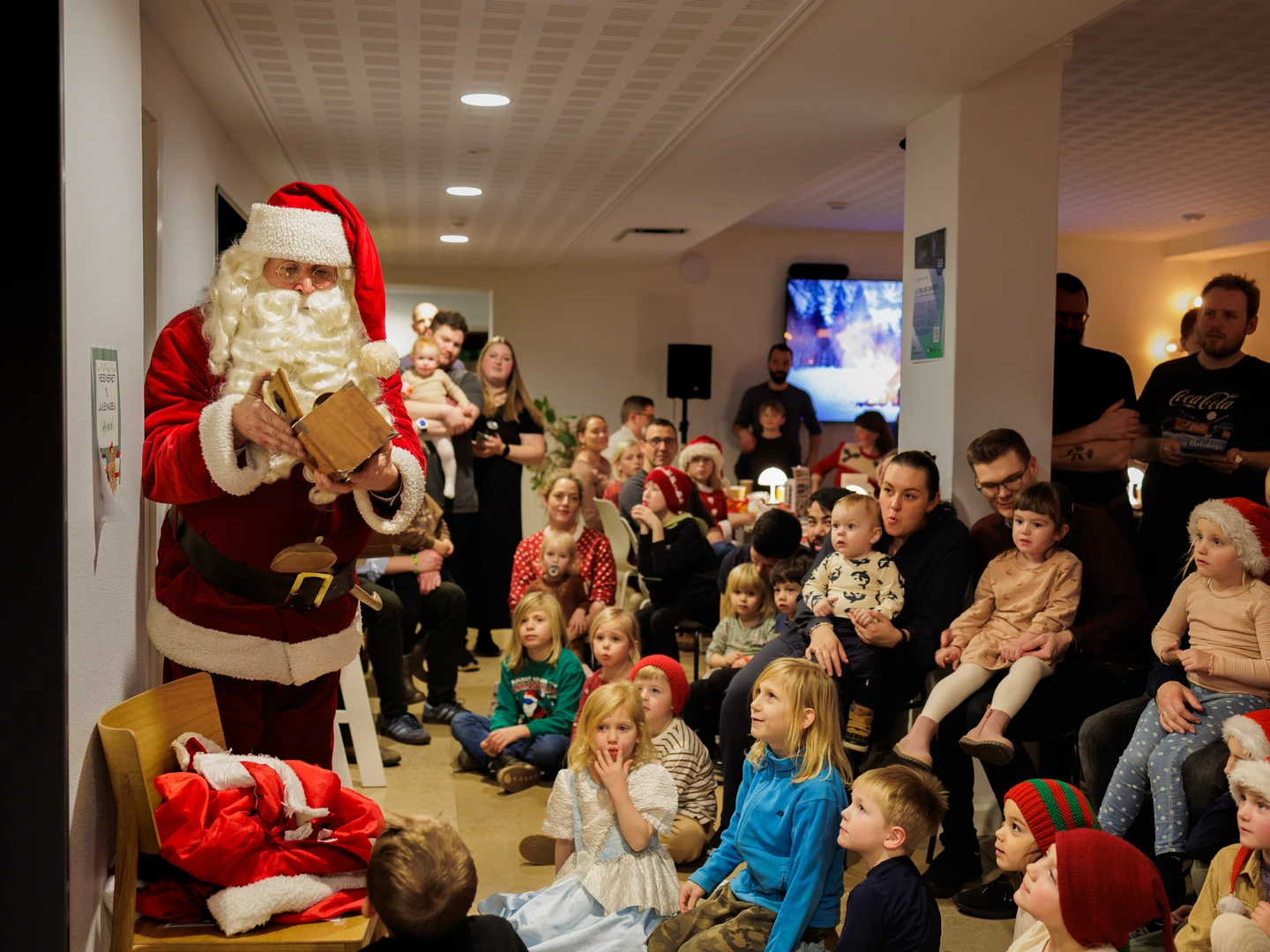 Julemanden deler gaver til en gruppe forventningsfulde børn og voksne i et festligt, indendørs rum, dekoreret til jul.