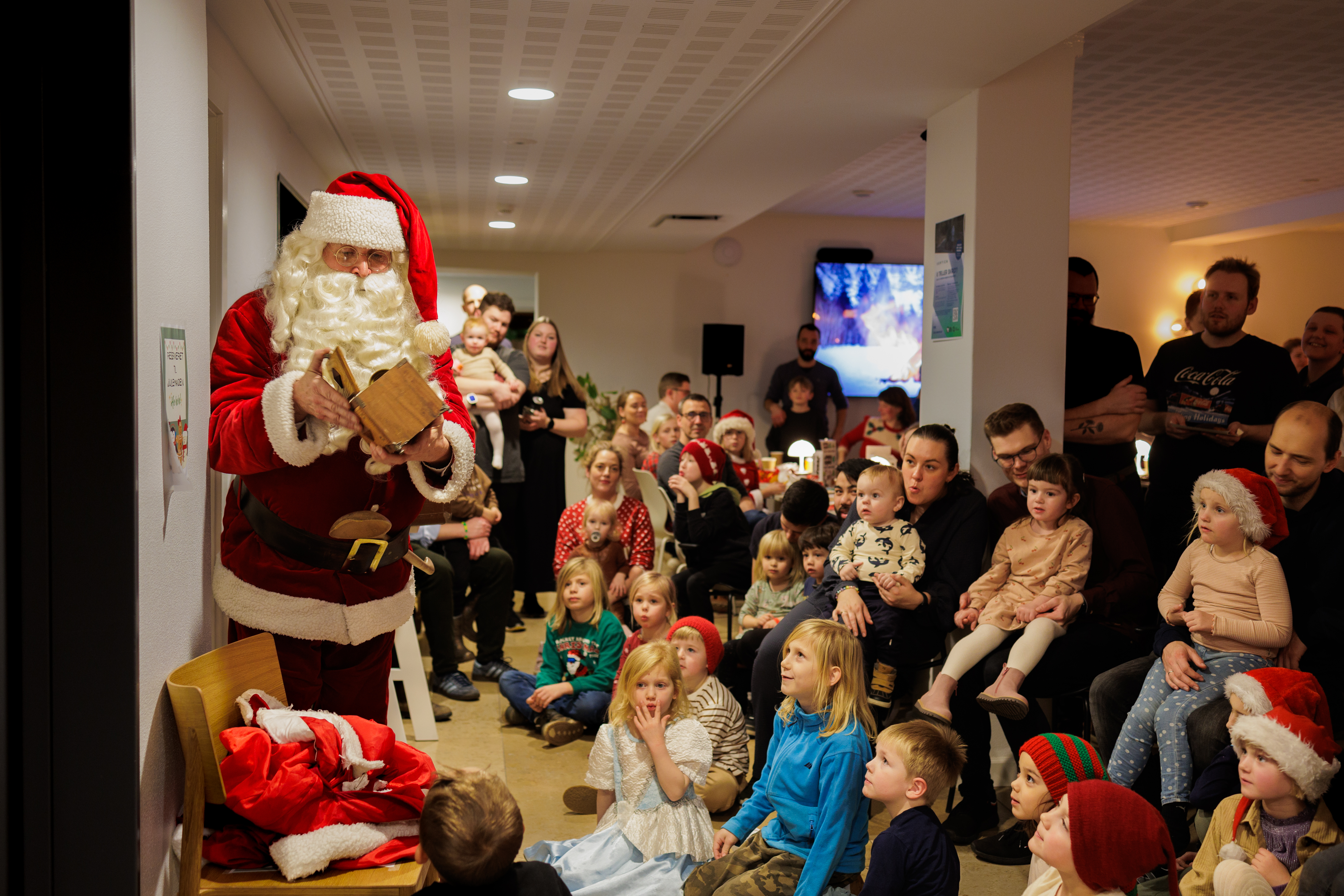 Julemanden deler gaver til en gruppe forventningsfulde børn og voksne i et festligt, indendørs rum, dekoreret til jul.