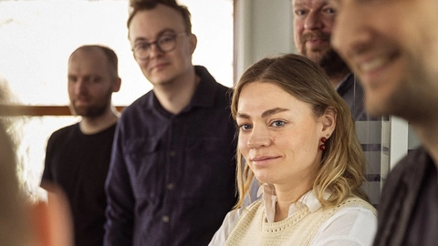 A group of people standing together in a bright room, with a woman in a knitted sweater and white blouse smiling thoughtfully while others look on.