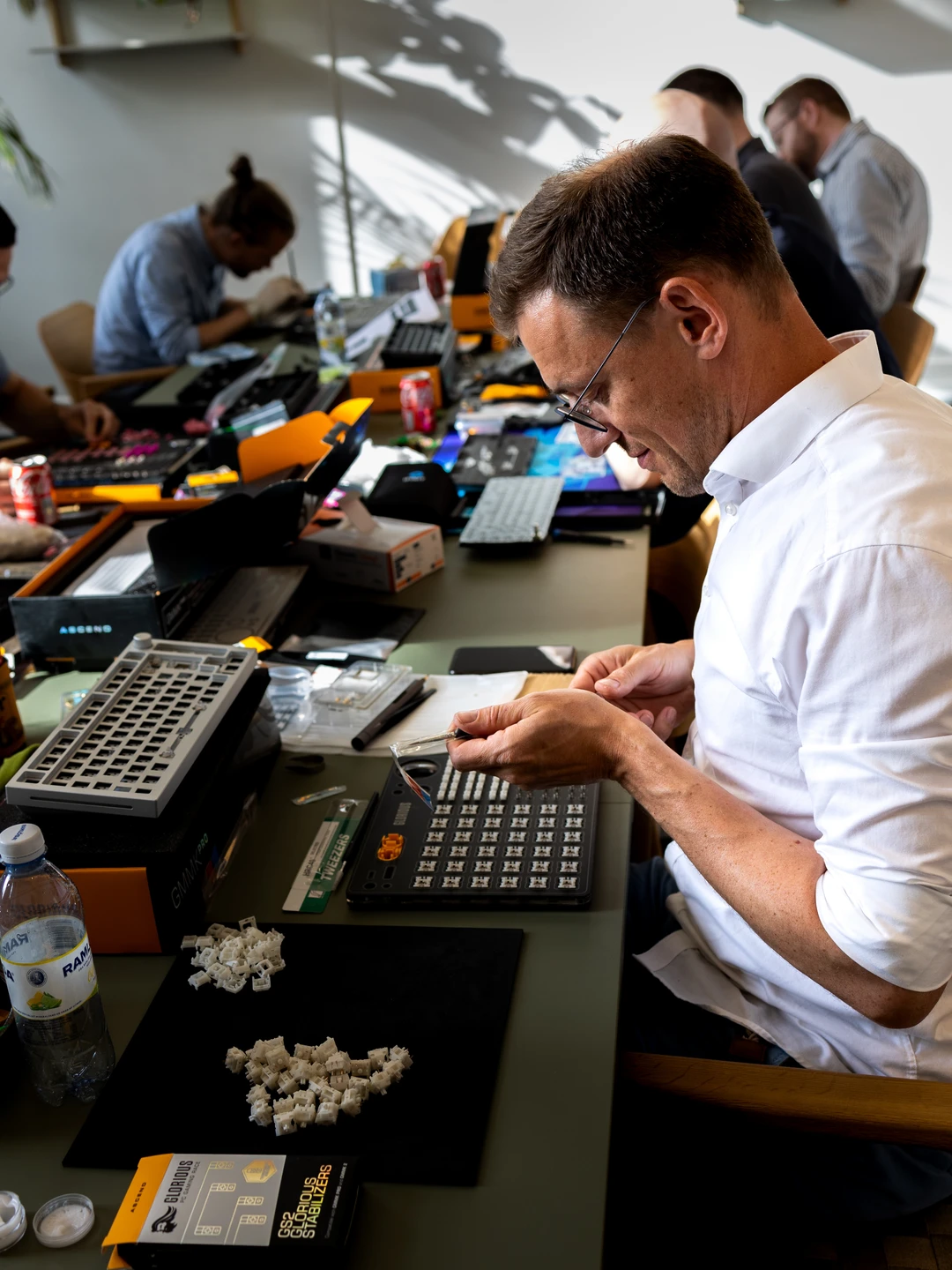 En mand iført en hvid skjorte arbejder koncentreret på at samle tastaturkomponenter ved et bord fyldt med diverse tastaturdele, værktøj og snacks, mens andre personer i baggrunden også arbejder på deres tastaturer.