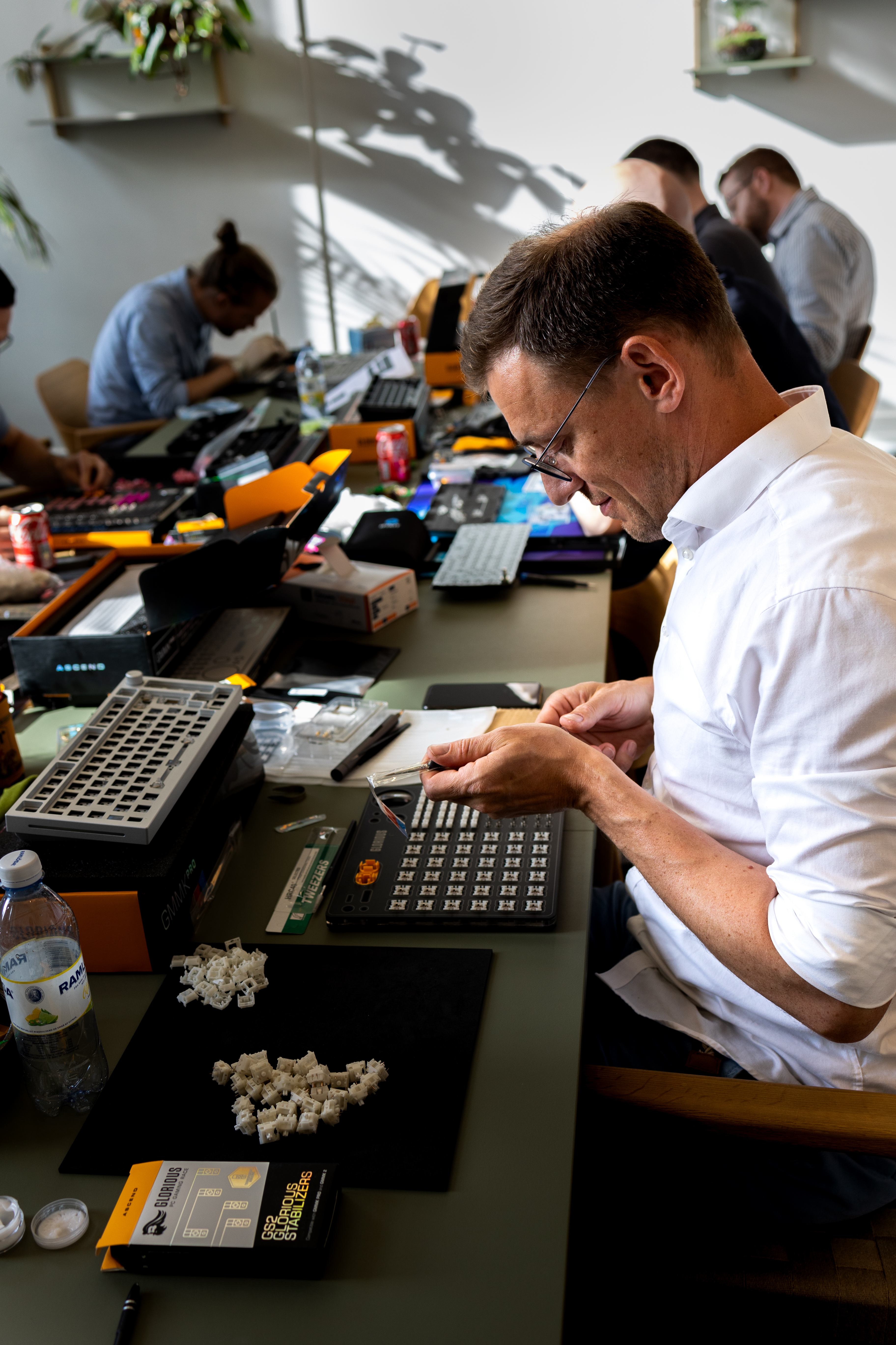 En mand iført en hvid skjorte arbejder koncentreret på at samle tastaturkomponenter ved et bord fyldt med diverse tastaturdele, værktøj og snacks, mens andre personer i baggrunden også arbejder på deres tastaturer.