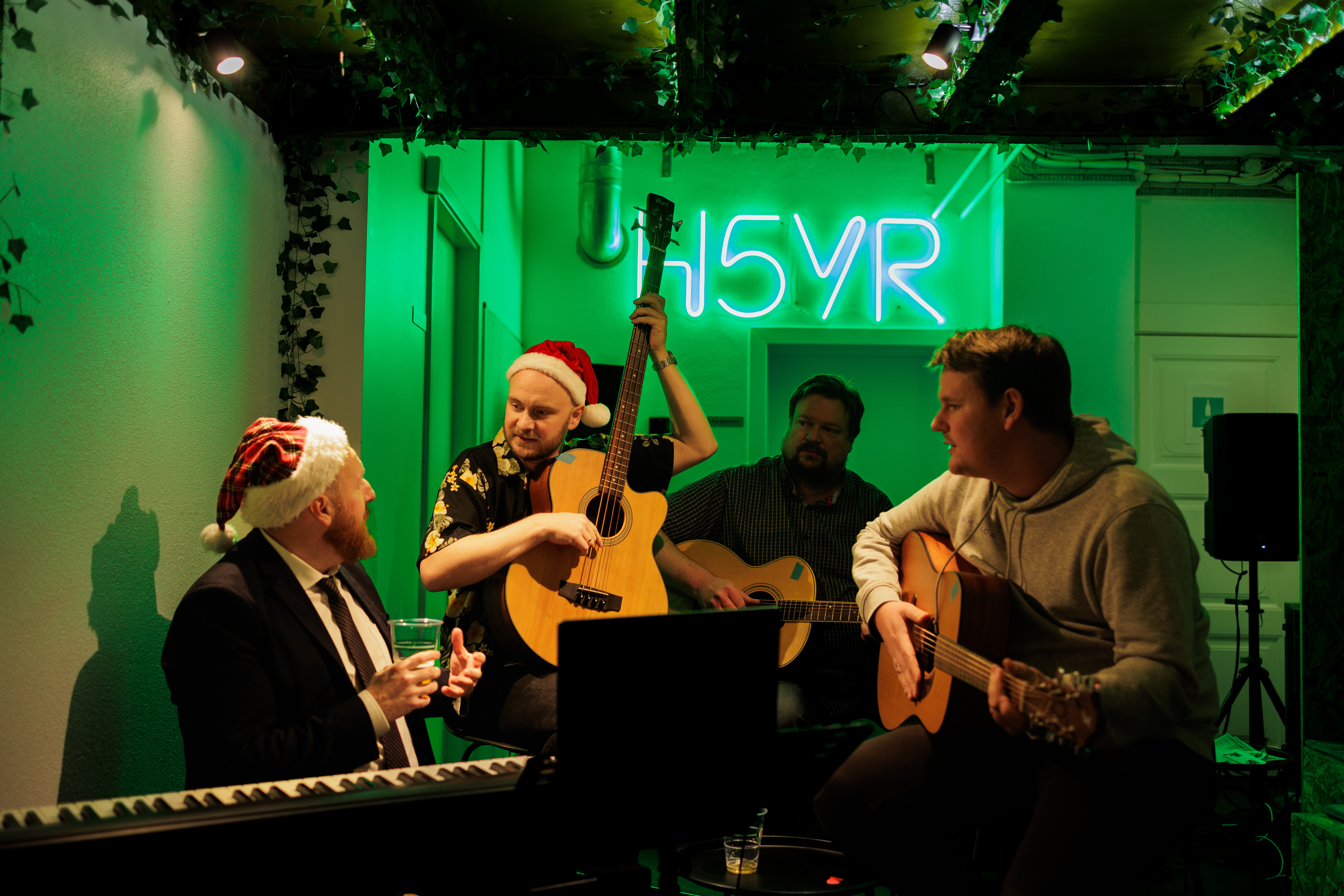 Fire mænd iført nissehuer sidder i et musikmiljø med grøn belysning, mens de spiller guitar og snakker. En mand i jakkesæt holder et glas, mens de nyder musikken i en afslappet atmosfære.