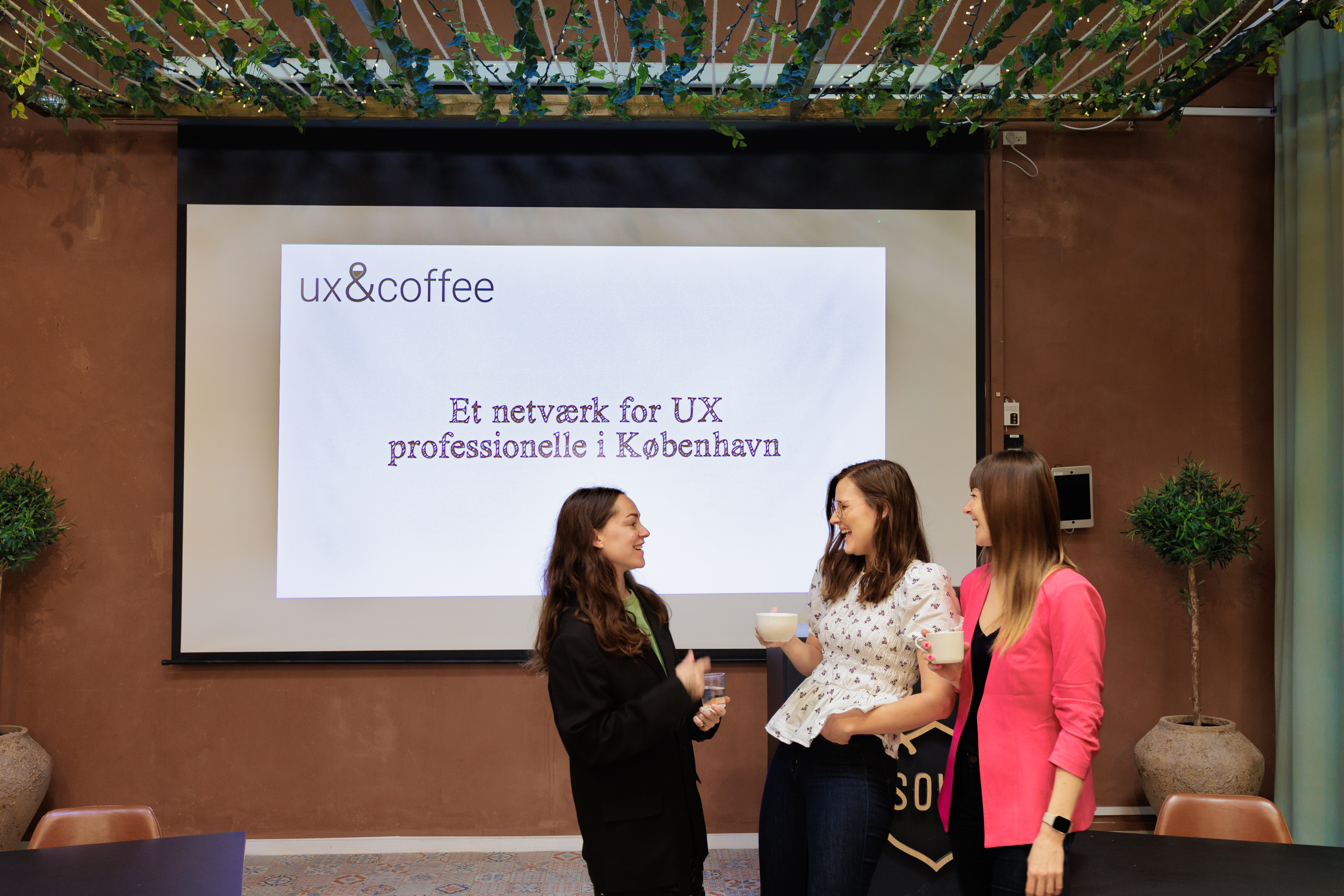3 kvinder står sammen og smiler eller griner stort, kvinderne står foran en skærm der viser teksten "Et netværk for UX professionelle i København"
