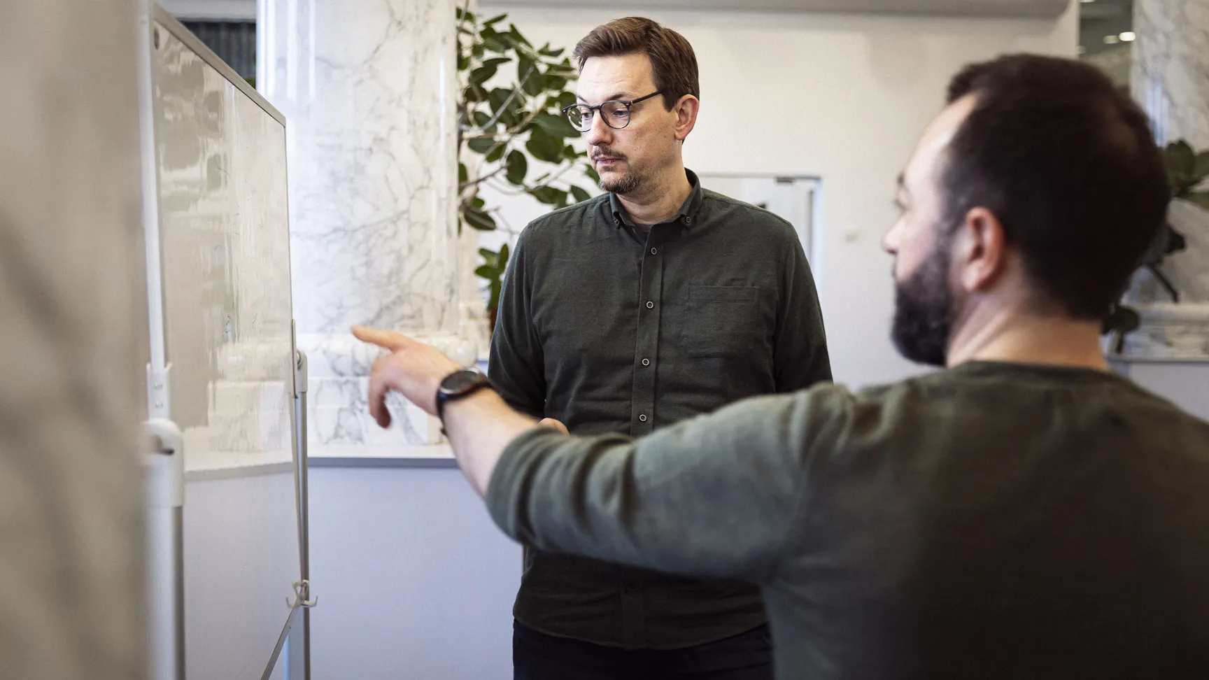 To mænd i en kontormiljø diskuterer over en whiteboard, hvor den ene mand peger og den anden ser opmærksomt på tavlen.