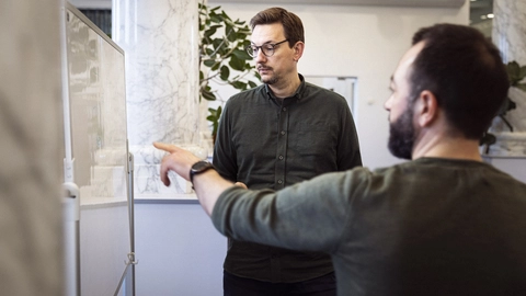 To mænd i en kontormiljø diskuterer over en whiteboard, hvor den ene mand peger og den anden ser opmærksomt på tavlen.