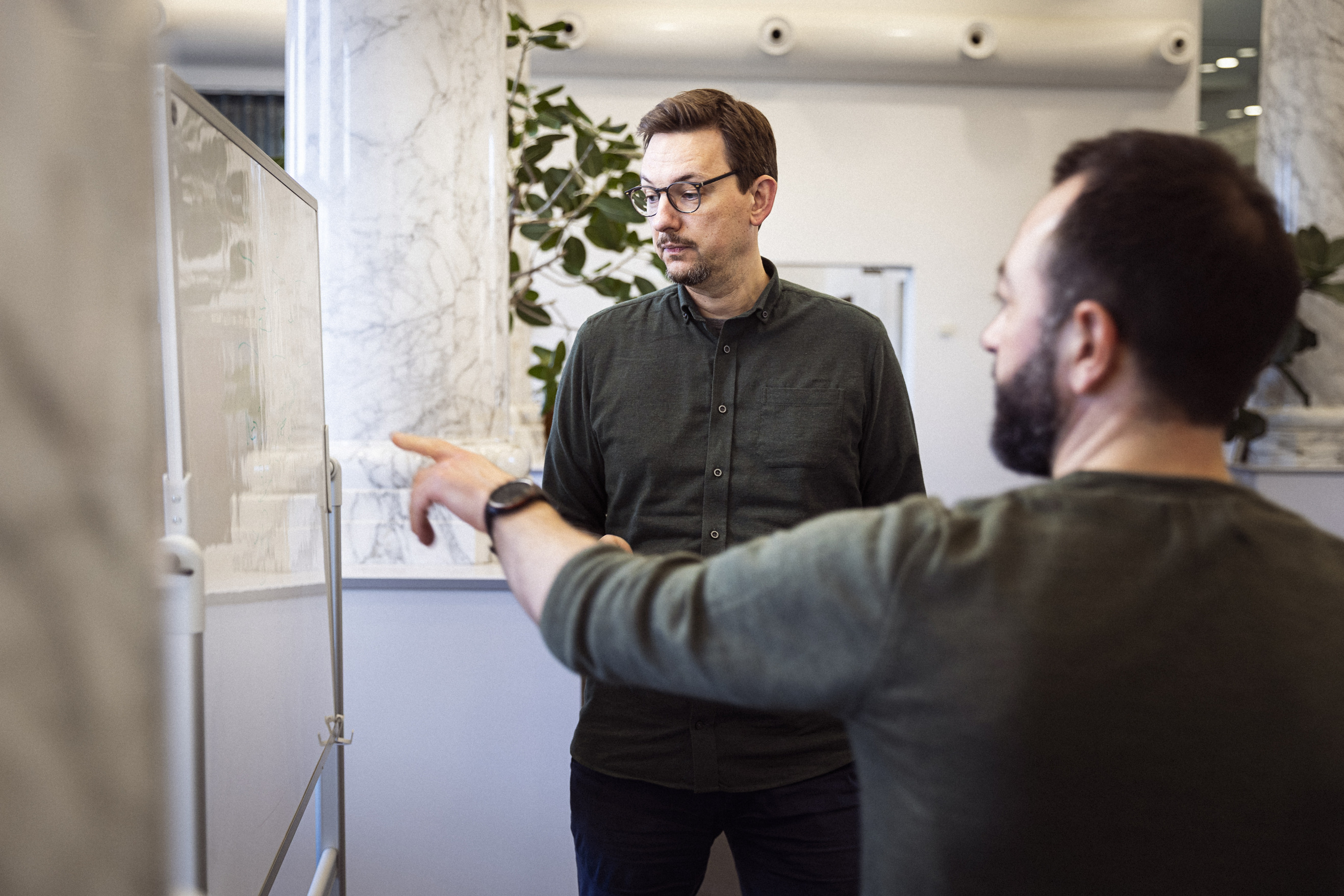 To mænd i en kontormiljø diskuterer over en whiteboard, hvor den ene mand peger og den anden ser opmærksomt på tavlen.