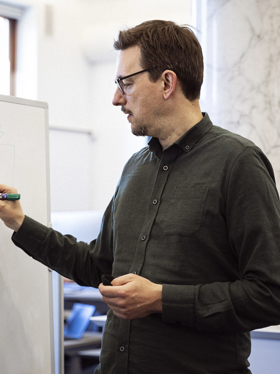 En mand står ved en tavle og tegner diagrammer med en grøn tusch, mens to personer til venstre ser interesseret til. Kontormiljø med moderne indretning i baggrunden.
