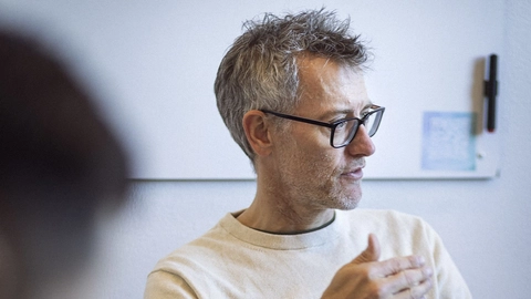 En ældre mand med gråt hår og briller sidder ved et bord og gestikulerer, mens han taler. Han bærer en lys trøje og der er en bærbar computer foran ham sammen med en vandflaske og en fjernbetjening. I baggrunden ses en hvid væg.
