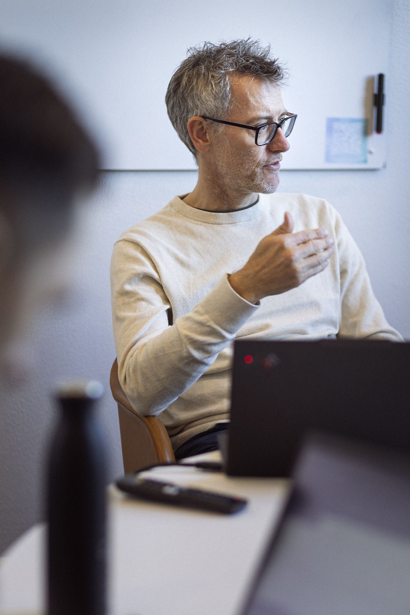 En ældre mand med gråt hår og briller sidder ved et bord og gestikulerer, mens han taler. Han bærer en lys trøje og der er en bærbar computer foran ham sammen med en vandflaske og en fjernbetjening. I baggrunden ses en hvid væg.