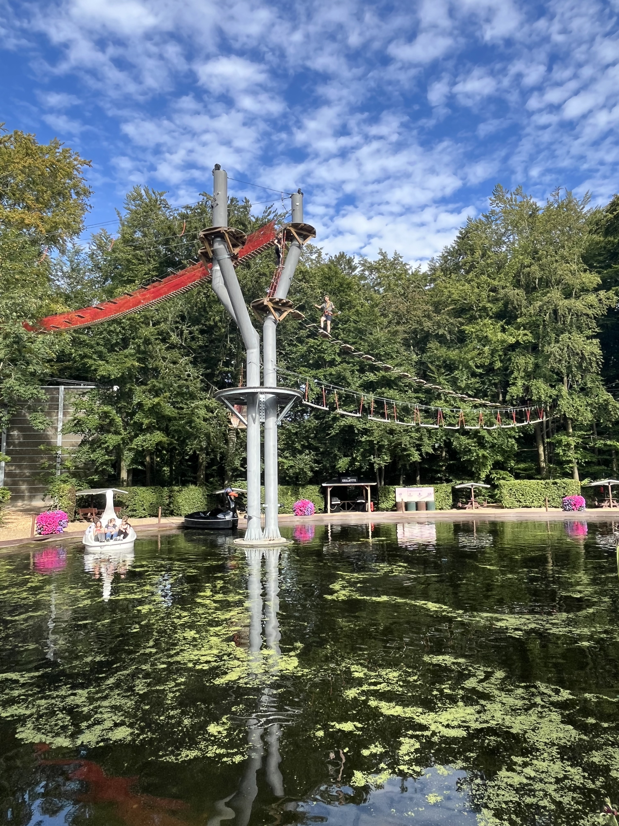 En person balancerer på en høj rebbane over en sø, omgivet af grønne træer og pink blomster, med en klar blå himmel i baggrunden.