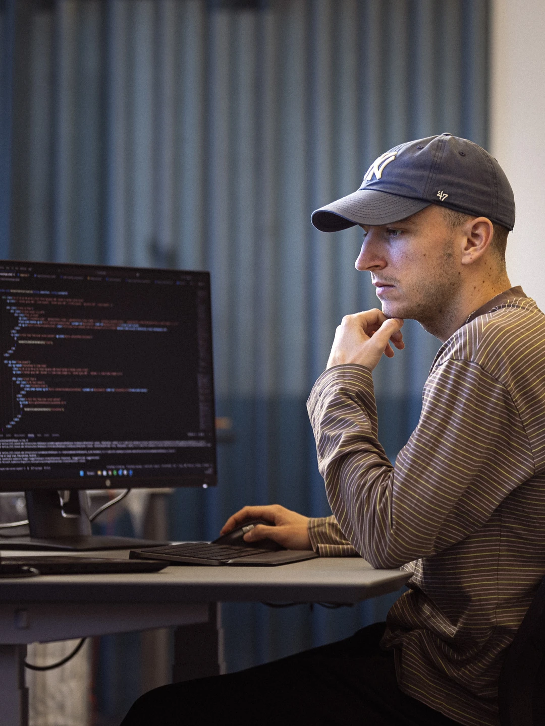 En mand i kasket arbejder koncentreret ved en computer med flere skærme, der viser programmeringskode.