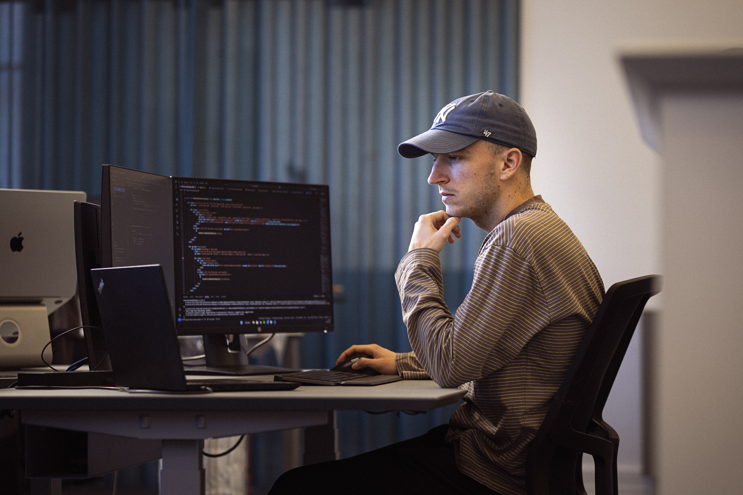 En mand i kasket arbejder koncentreret ved en computer med flere skærme, der viser programmeringskode.
