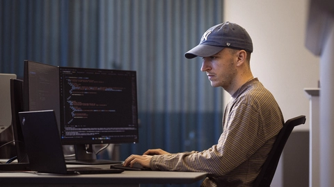 En mand i kasket arbejder koncentreret ved en computer med flere skærme, der viser programmeringskode.