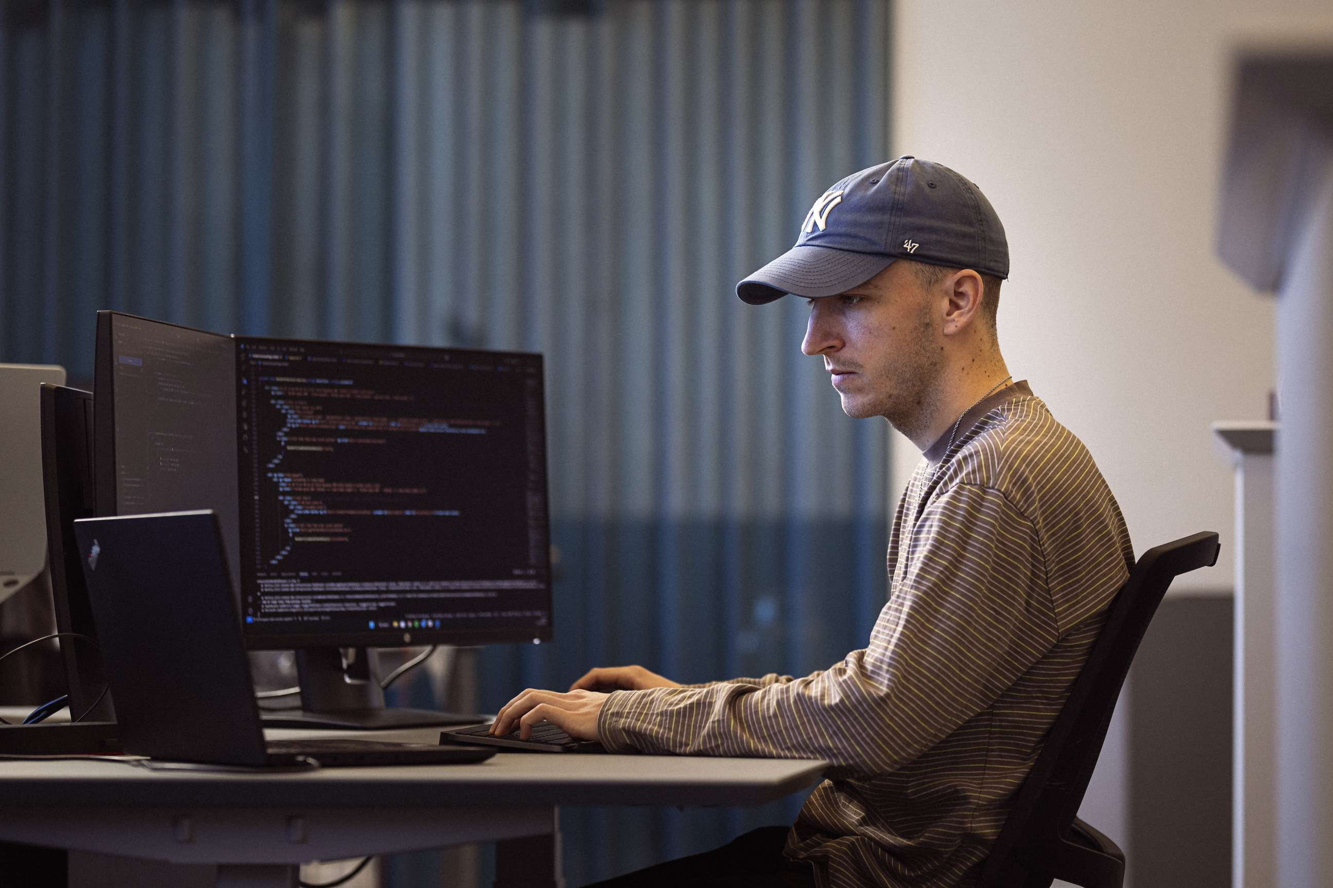 En mand i kasket arbejder koncentreret ved en computer med flere skærme, der viser programmeringskode.