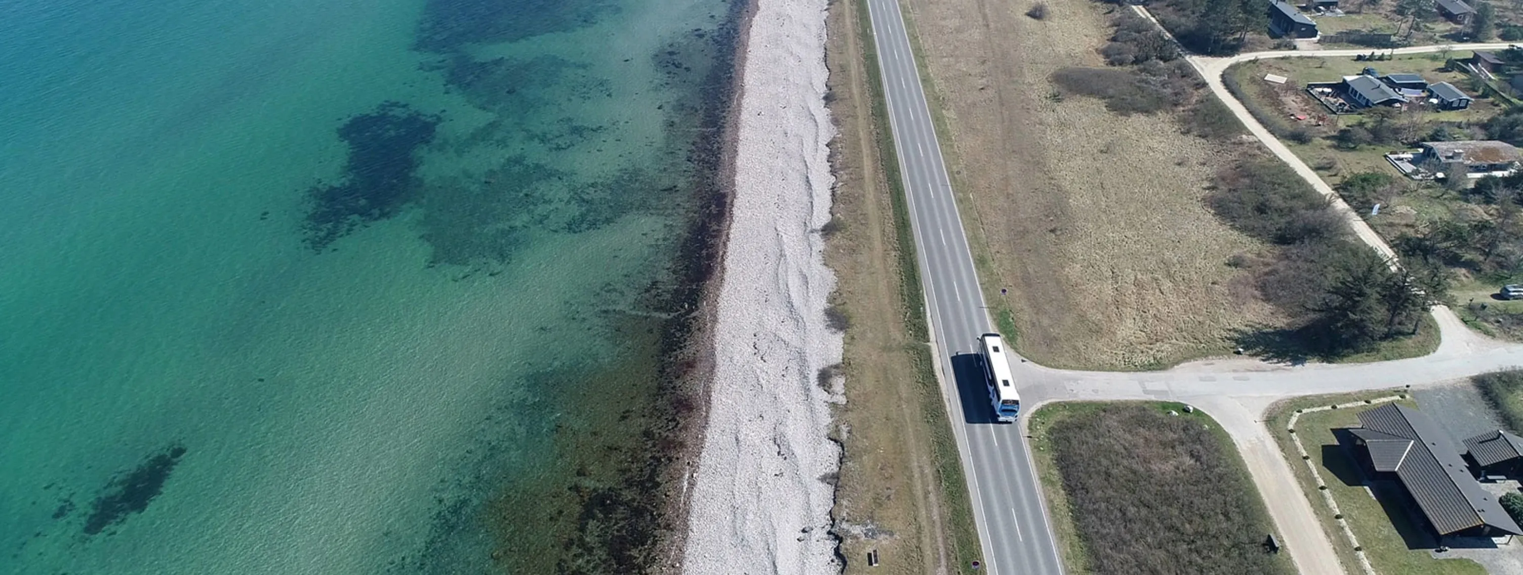 Oversigt over en strandlinje med klart blåt vand, sten og græsarealer ved siden af en vej, hvor en bus kører forbi.