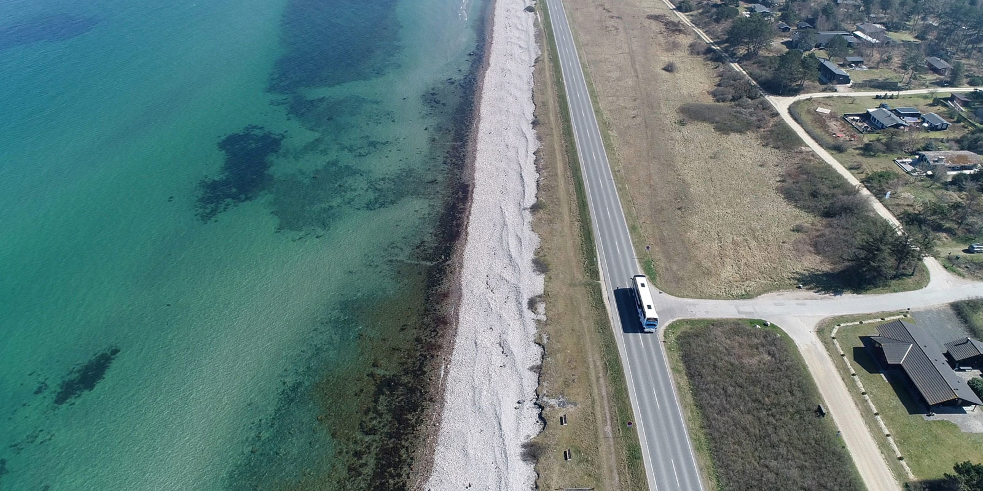 Oversigt over en strandlinje med klart blåt vand, sten og græsarealer ved siden af en vej, hvor en bus kører forbi.