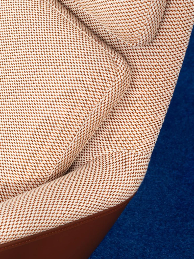 Close-up of a modern upholstered chair featuring a textured beige and brown fabric, highlighting its curved shape and soft cushioning against a blue background.