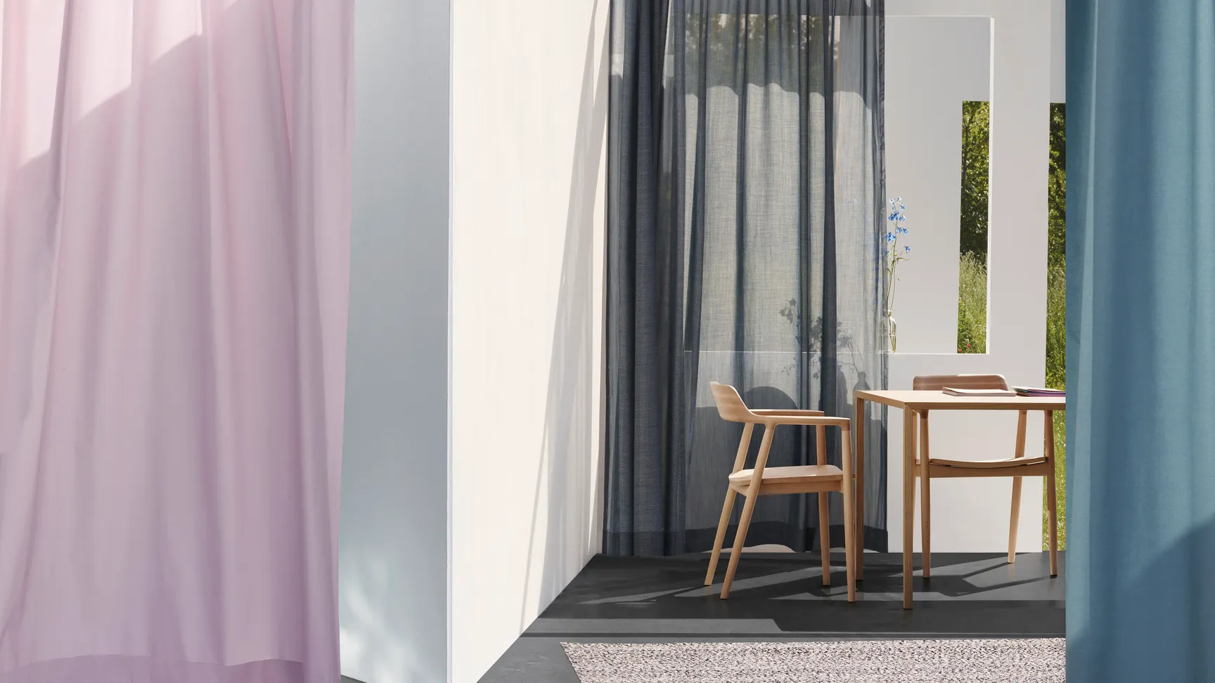 A cozy interior space featuring sheer curtains in soft pink and dark blue, a wooden dining table with two chairs, a neutral rug, and natural light streaming through a window, enhancing the ambiance.