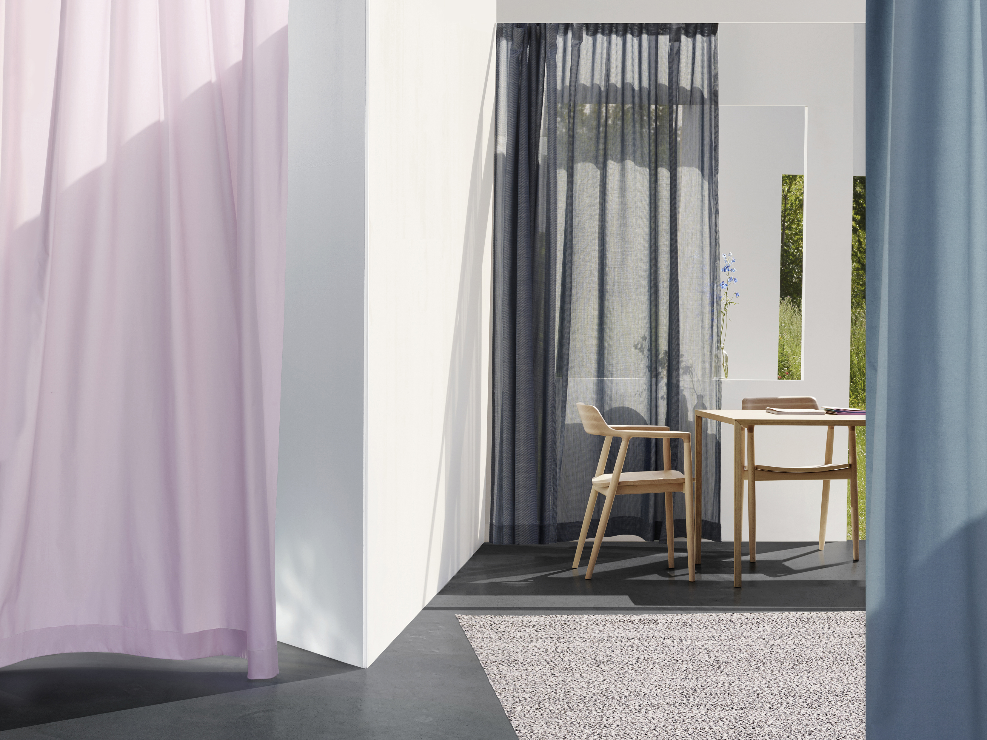 A cozy interior space featuring sheer curtains in soft pink and dark blue, a wooden dining table with two chairs, a neutral rug, and natural light streaming through a window, enhancing the ambiance.