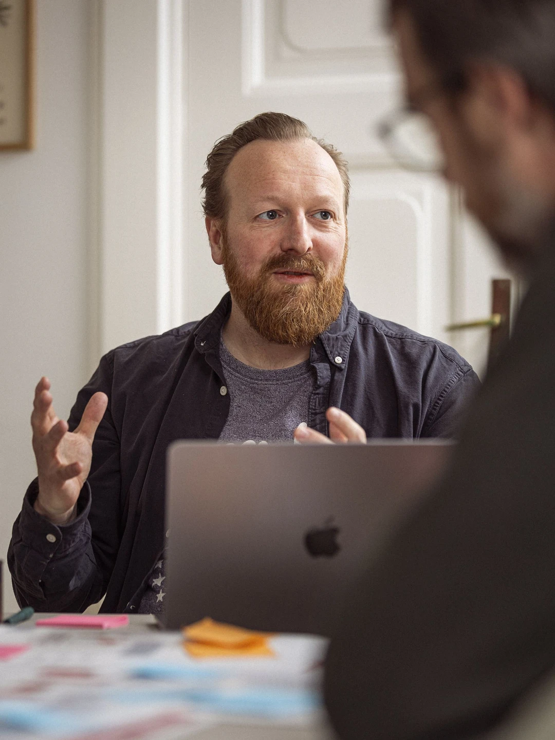 Mand med skæg og brunt hår sidder ved et skrivebord og taler, mens han gestikulerer med hænderne. I baggrunden ses en anden person delvist i fokus og en bærbar computer. Der er farverige post-its på bordet, som bidrager til en kreativ arbejdsstemning.