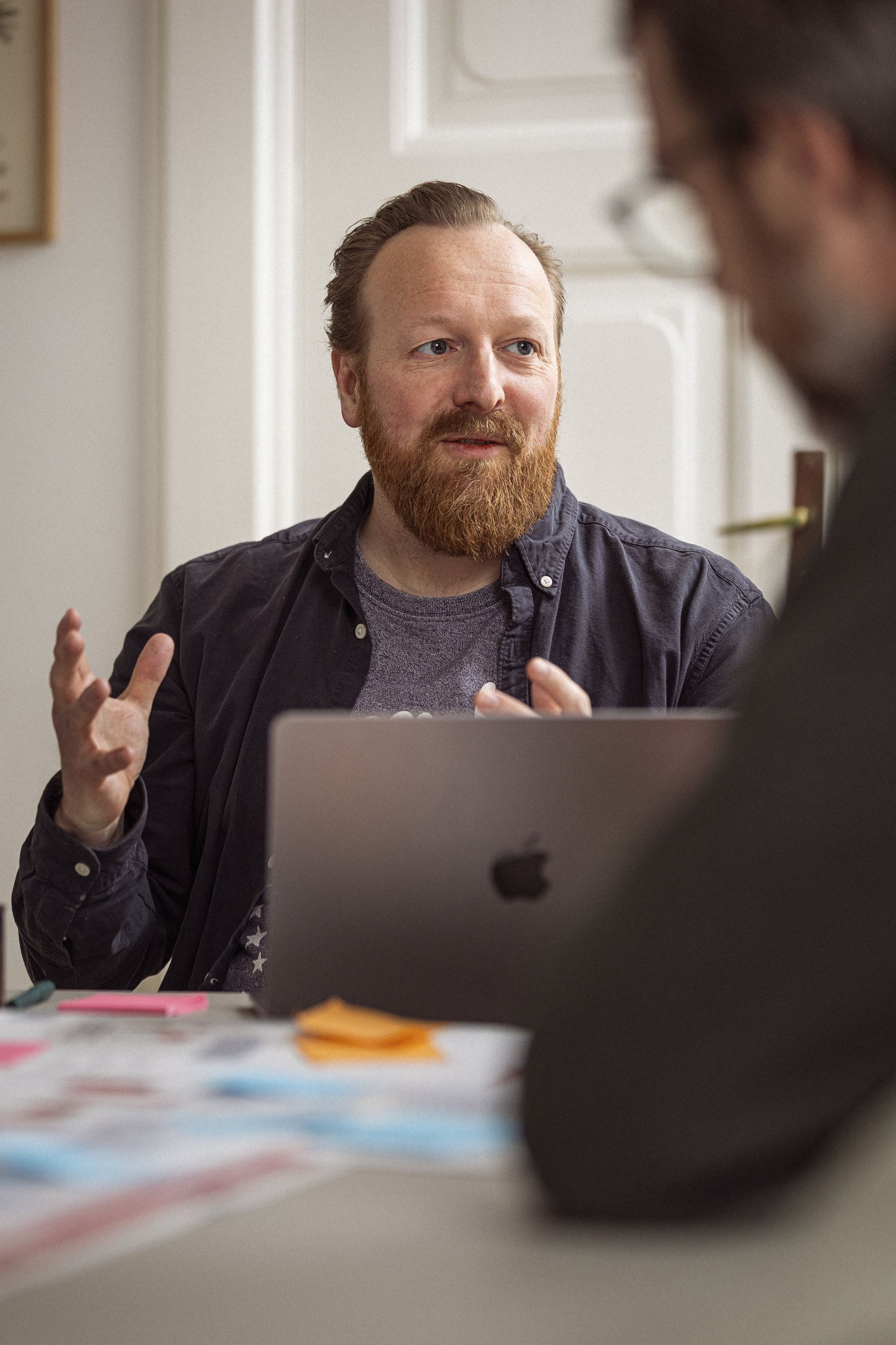 Mand med skæg og brunt hår sidder ved et skrivebord og taler, mens han gestikulerer med hænderne. I baggrunden ses en anden person delvist i fokus og en bærbar computer. Der er farverige post-its på bordet, som bidrager til en kreativ arbejdsstemning.