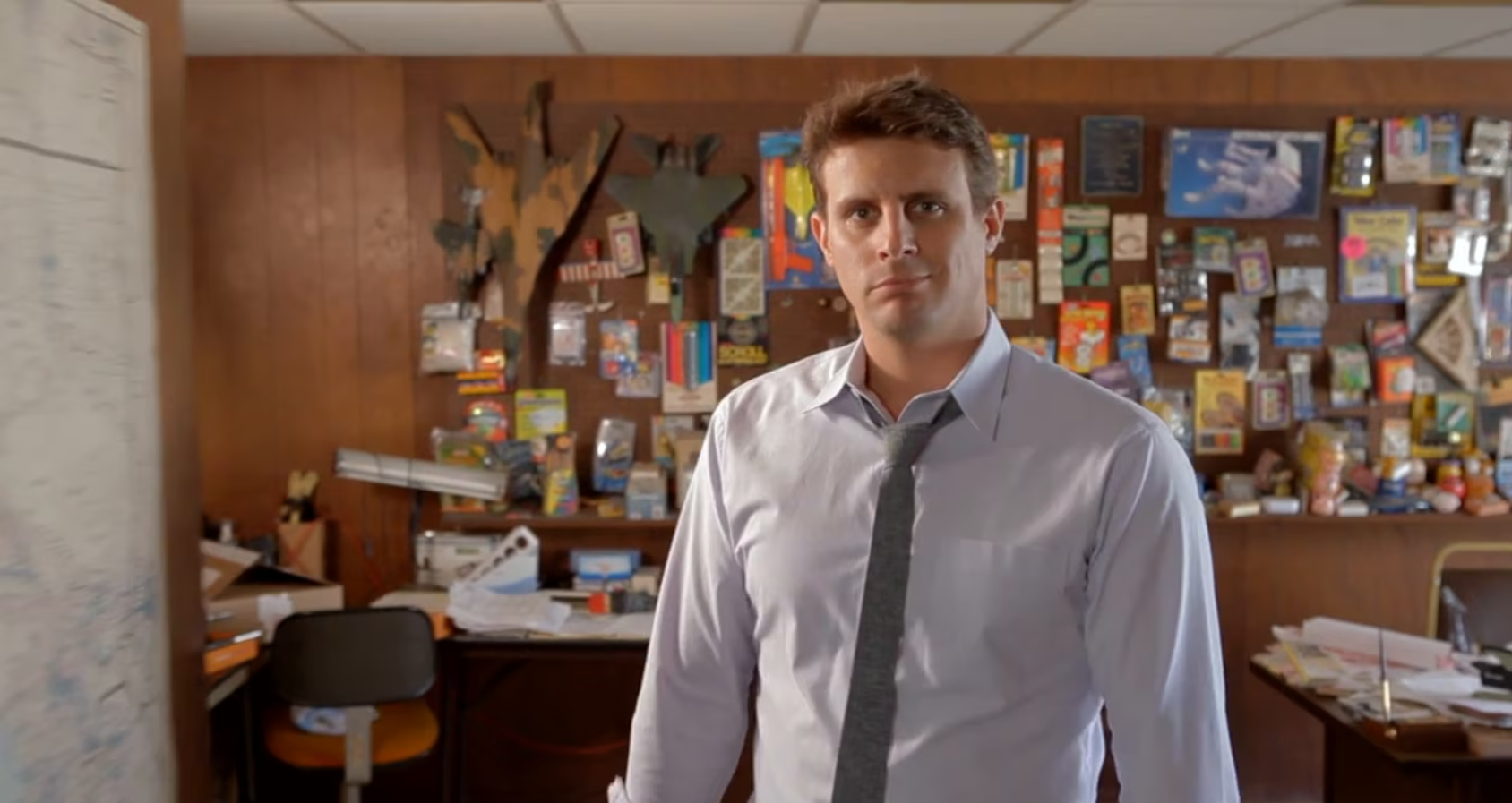 Mand i skjorte og slips står i et kontor med brune vægge, omgivet af farverige souvenirs og kontorartikler.