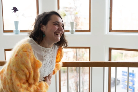 Ung kvinde med brunt hår iført en strikket orange cardigan, står ved et vindue med trærammer og smiler stort, mens hun holder sig til rækværket i en lys stue.